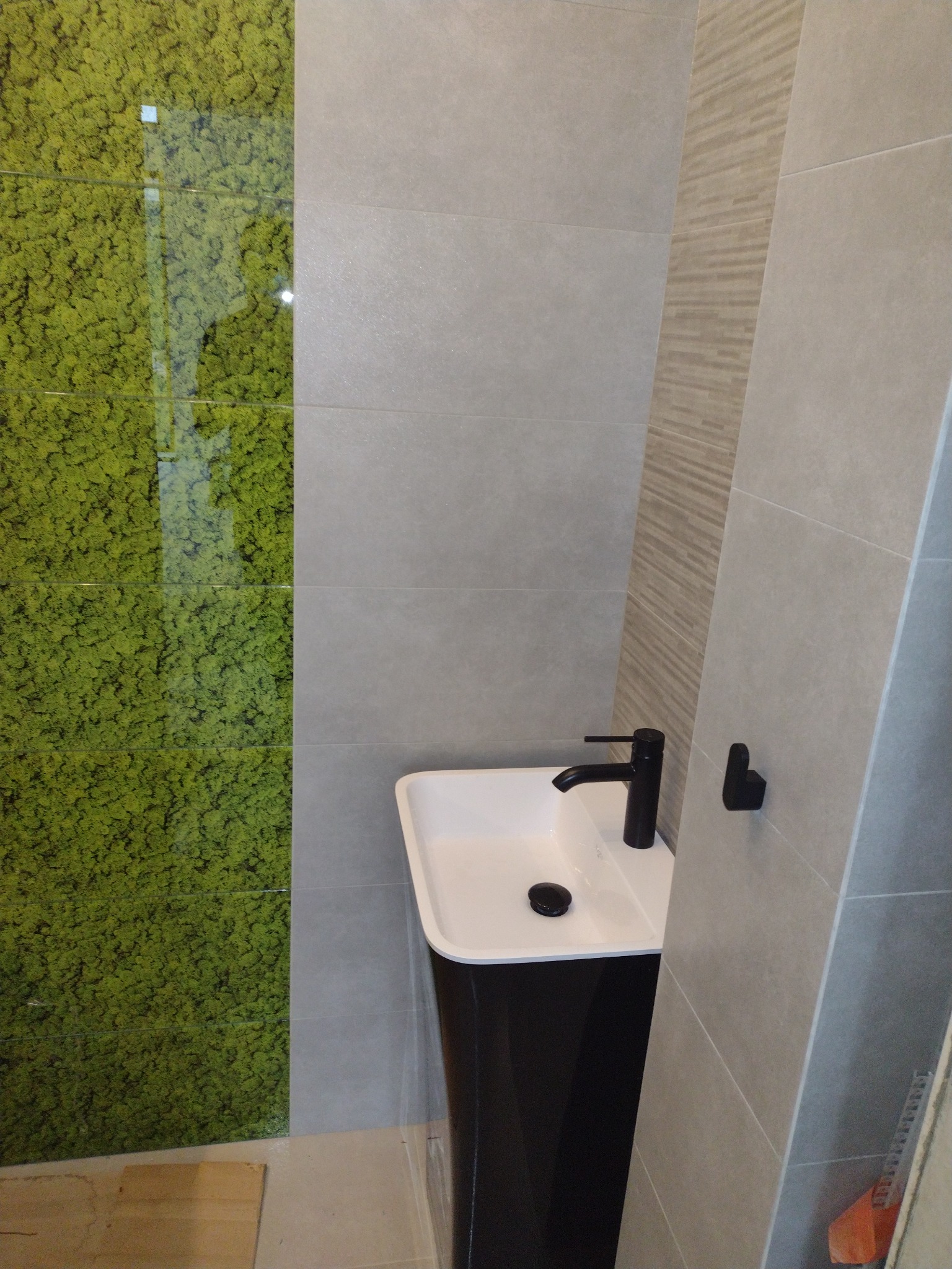 Nowoczesna łazienka z umywalką na czarnej nodze, czarną armaturą i ścianą wyłożoną szarymi płytkami oraz panelem z motywem zielonego mchu.