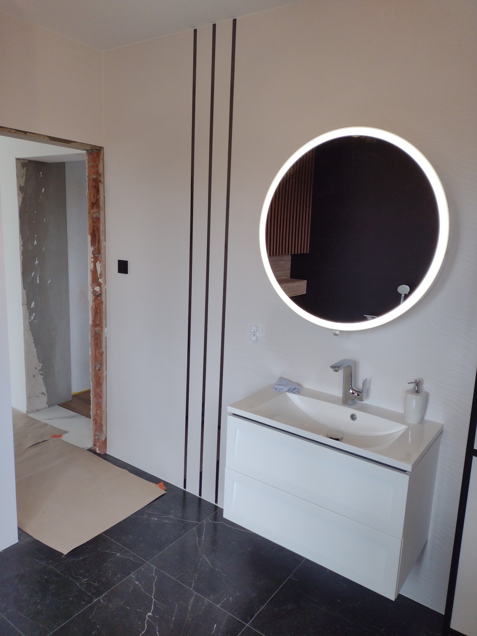 Nowoczesna łazienka w trakcie wykańczania: biała szafka z umywalką i okrągłym lustrem z podświetleniem LED, czarne płytki podłogowe z białym żyłkowaniem, ściana z pionowymi czarnymi pasami...