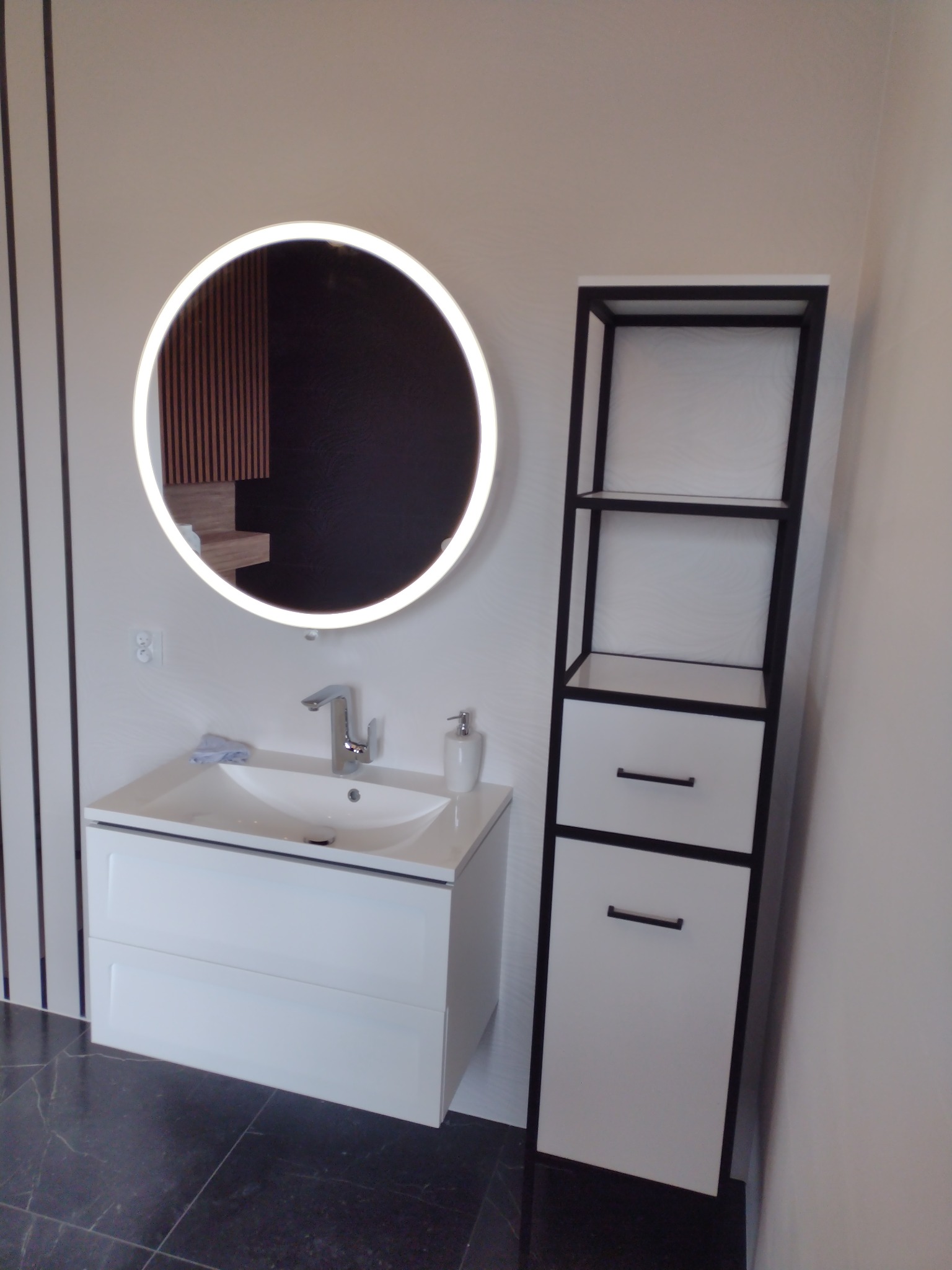 Nowoczesna łazienka: biała szafka pod umywalkę z okrągłym lustrem LED i czarno-biały regał na tle jasnych ścian i ciemnej podłogi.