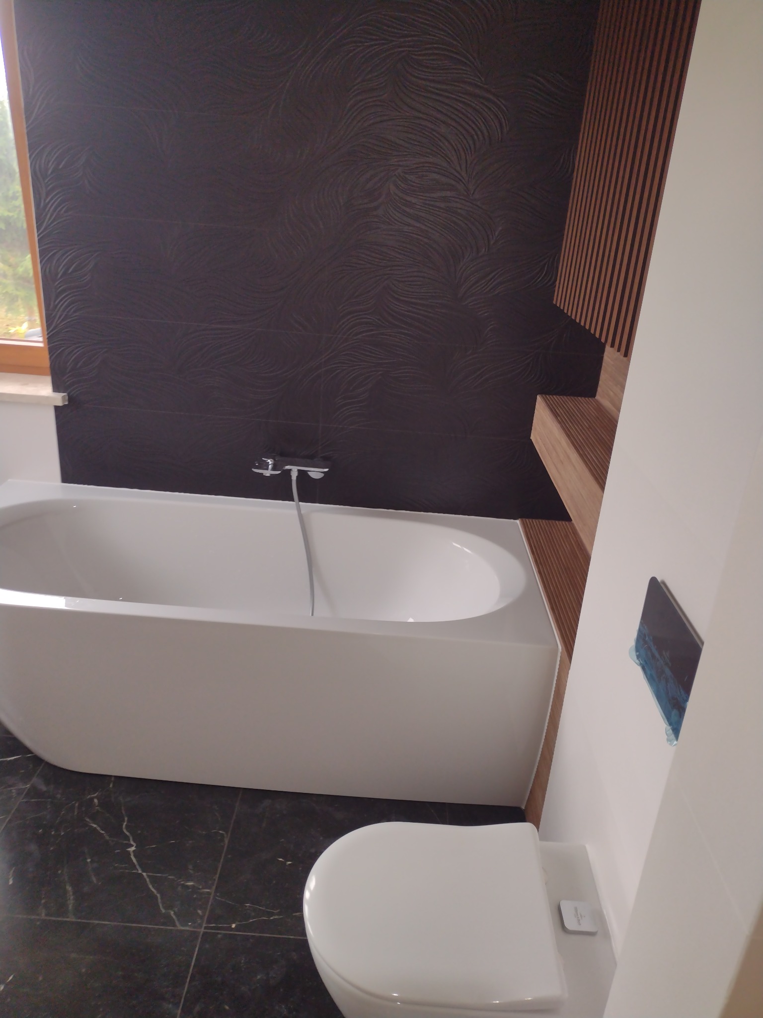 Nowoczesna łazienka z czarną, wzorzystą ścianą, białą wanną i toaletą, drewnianymi półkami i marmurową podłogą.