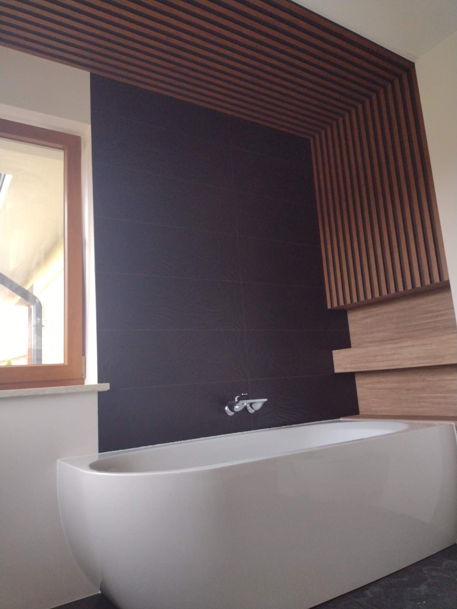 Nowoczesna łazienka z wolnostojącą białą wanną, ciemnymi płytkami na ścianie i drewnianą dekoracją sufitu oraz ściany.