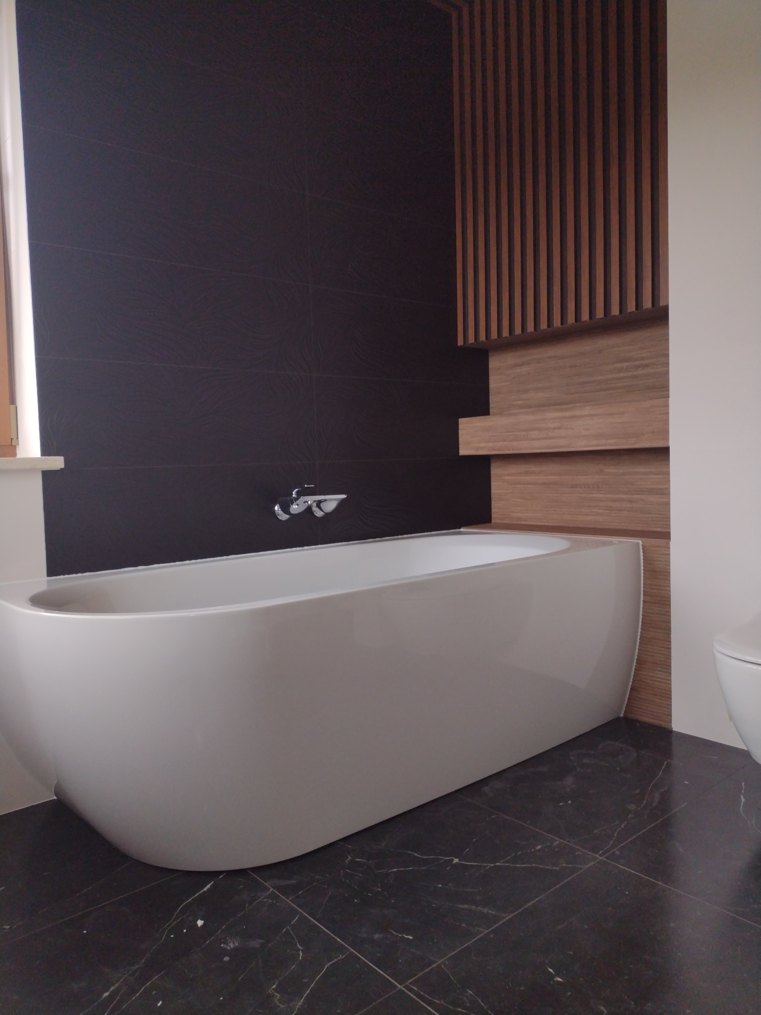 Nowoczesna łazienka z wolnostojącą białą wanną, ciemnymi płytkami na ścianie i podłodze imitującymi marmur oraz dekoracyjnym panelem z drewnianych listew.