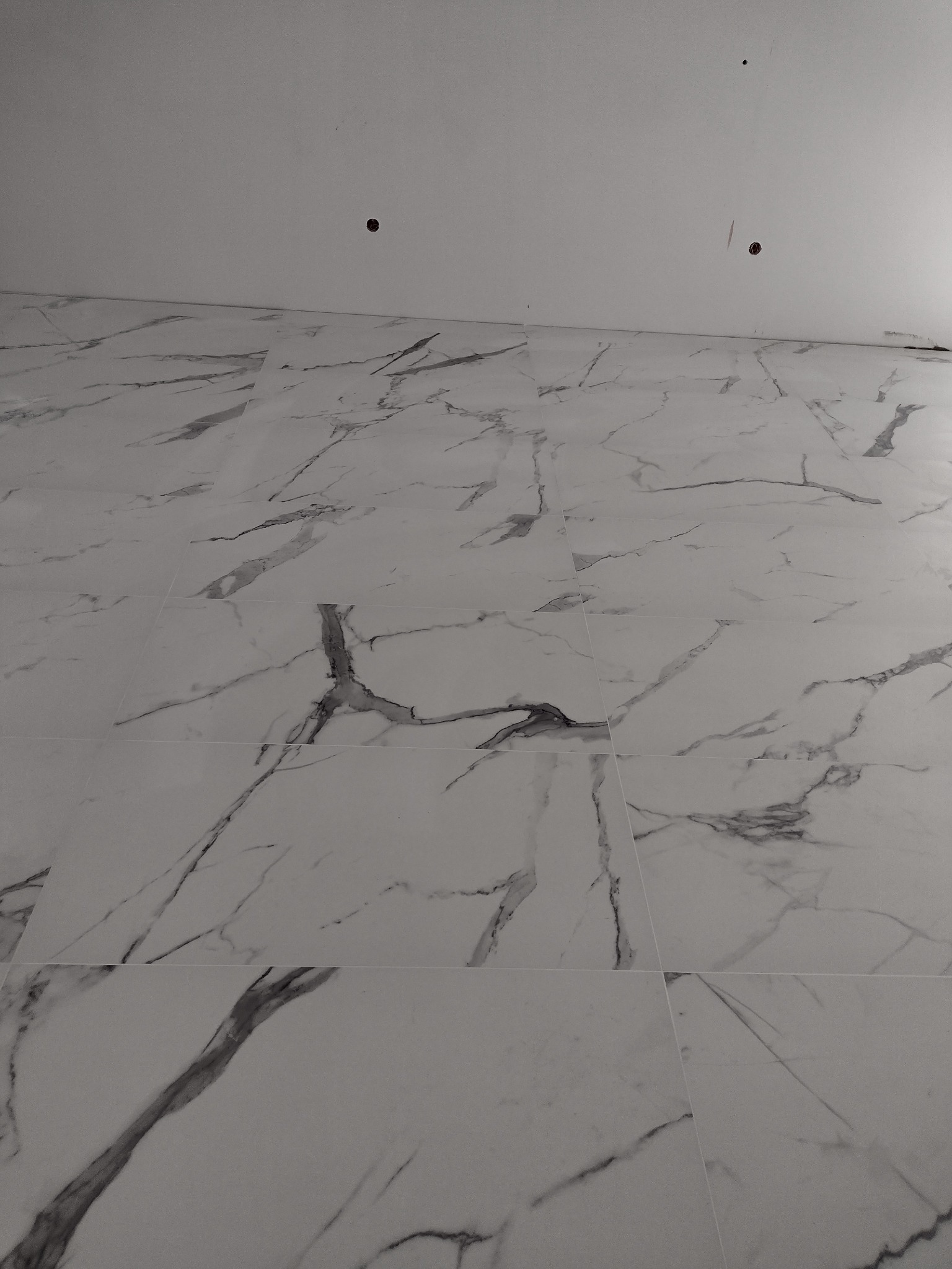 Świeżo ułożone, białe płytki imitujące marmur na ścianie łazienki, widoczne otwory w suficie pod oświetlenie.