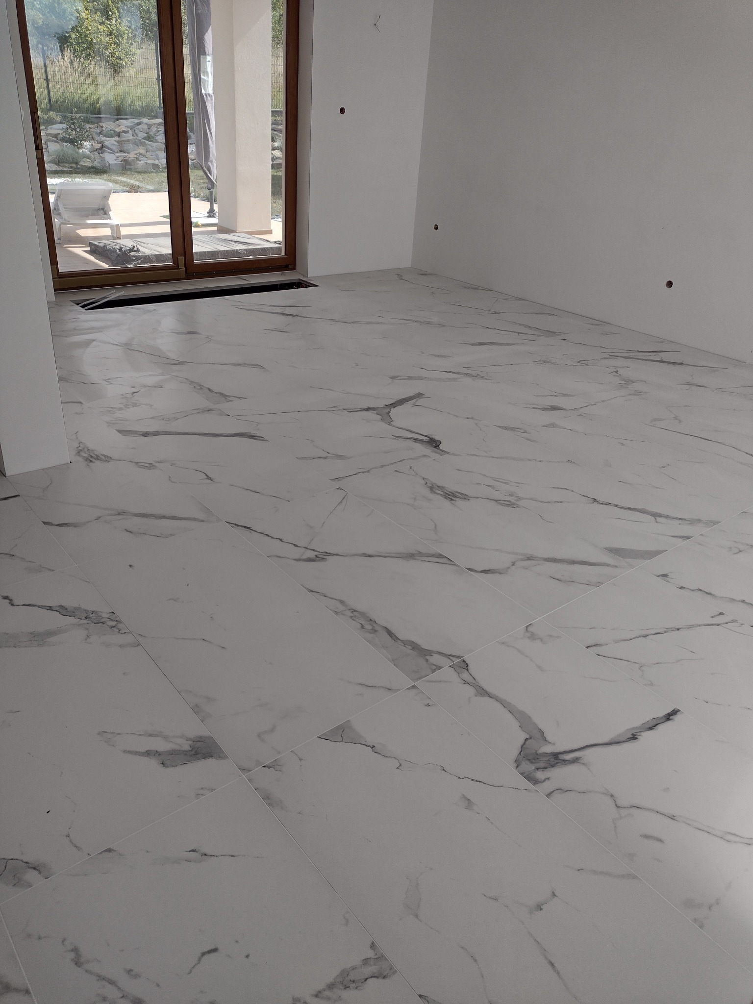 Jasna podłoga wyłożona białymi płytkami imitującymi marmur, widok na okno balkonowe z widokiem na ogród, surowe ściany gotowe do dalszych prac wykończeniowych.