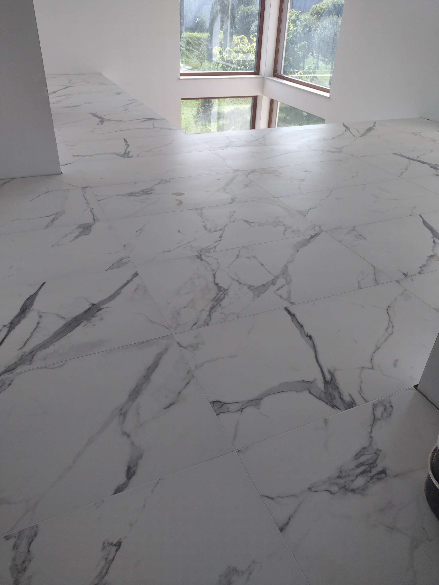 Białe płytki podłogowe imitujące marmur z widoczną fakturą na tle okna z widokiem na zieleń.