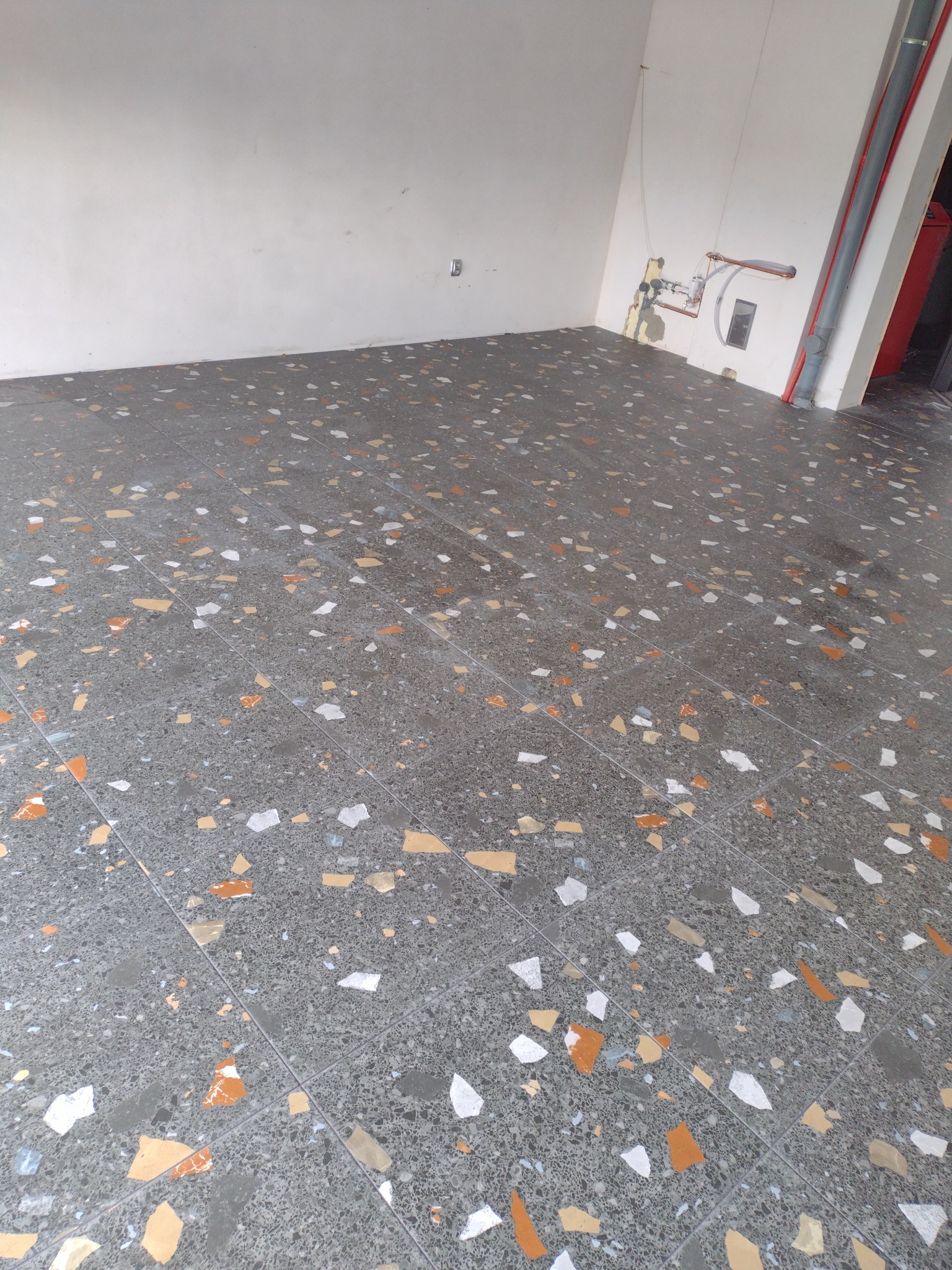 Szara posadzka z terazzo z białymi, pomarańczowymi i beżowymi fragmentami, widoczne rury i instalacja na ścianie w rogu pomieszczenia.