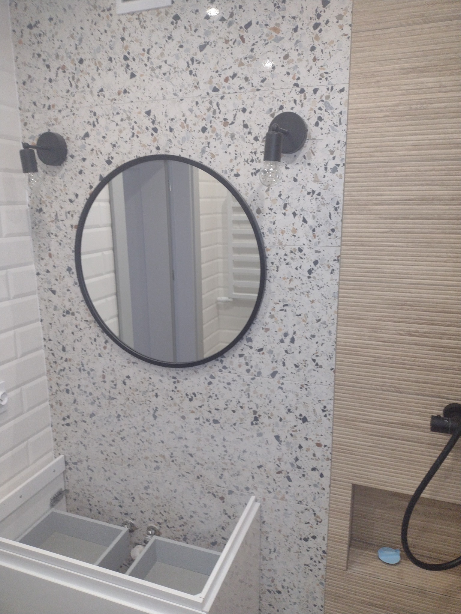 Nowoczesna łazienka z umywalką, okrągłym lustrem w czarnej ramie, dwoma czarnymi lampami ściennymi i płytkami w stylu lastryko na ścianie obok płytek imitujących drewno.