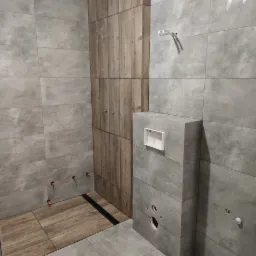 Wnętrze łazienki w trakcie remontu, z szarymi płytkami imitującymi beton, drewnopodobnymi płytkami na podłodze, zabudową z drewnopodobnych paneli i widocznymi elementami instalacji hydraulicznej.