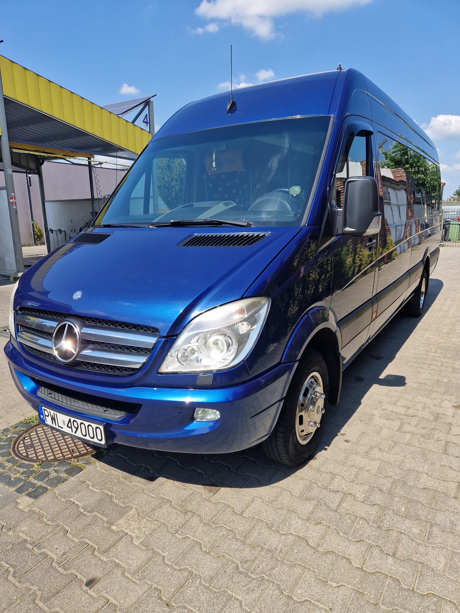 Niebieski bus Mercedes-Benz Sprinter z chromowanymi felgami i anteną na dachu, zaparkowany na kostce brukowej przed myjnią samochodową w słoneczny dzień.
