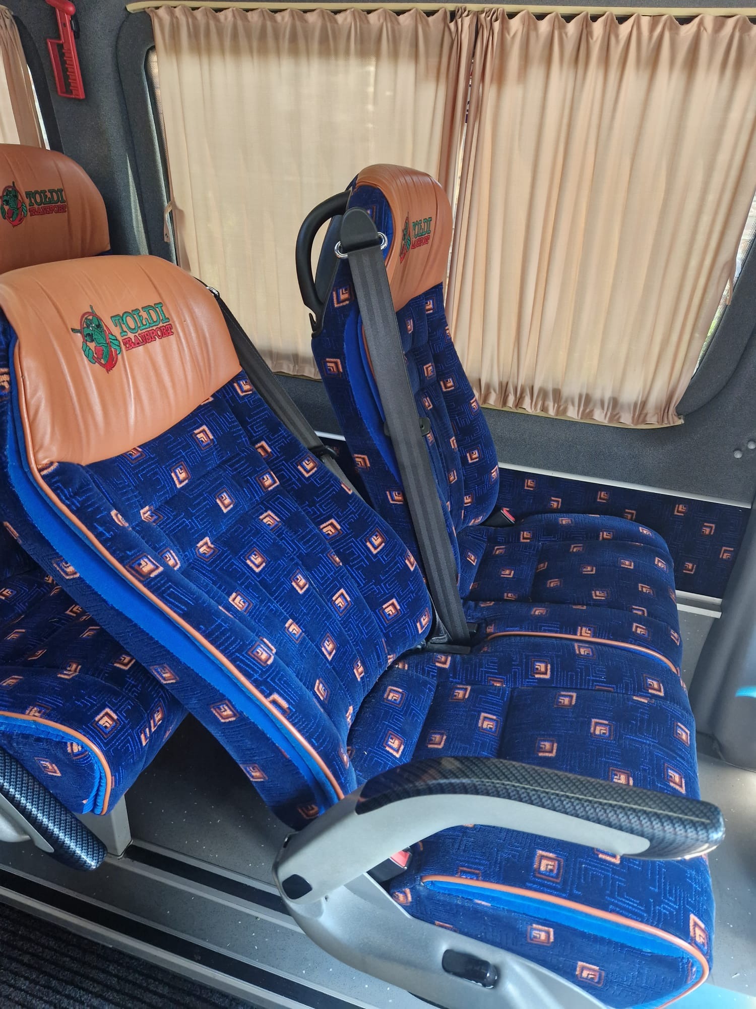 Wnętrze autobusu turystycznego z niebieskimi siedzeniami w geometryczne wzory i brązowymi zagłówkami z logo firmy transportowej, widok z boku na pasy bezpieczeństwa i zasłonięte okna.