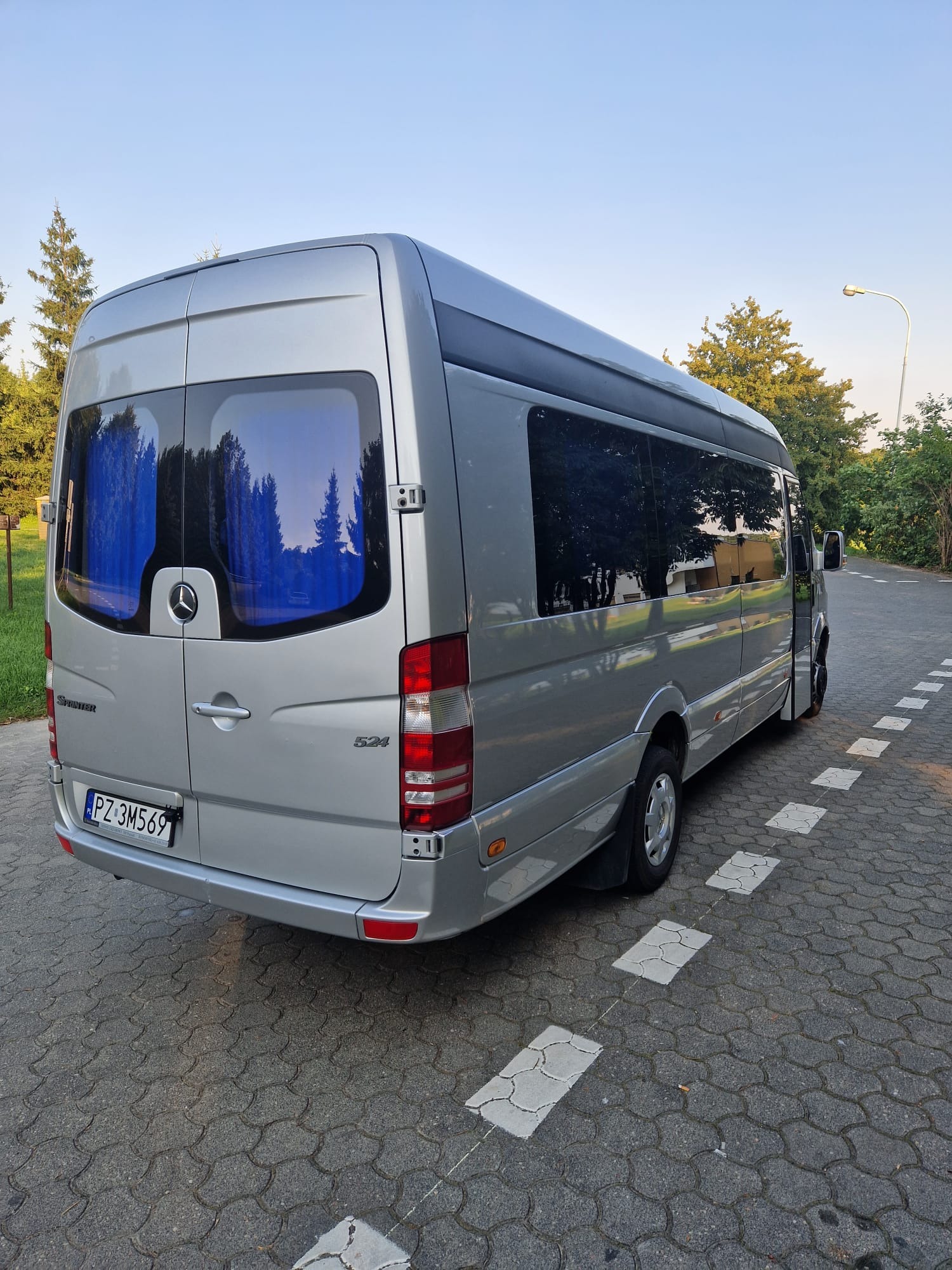 Srebrny bus Mercedes Sprinter 524 z ciemnymi szybami, widok z tyłu na kostce brukowej z namalowanymi białymi kwadratami, drzewa w tle.