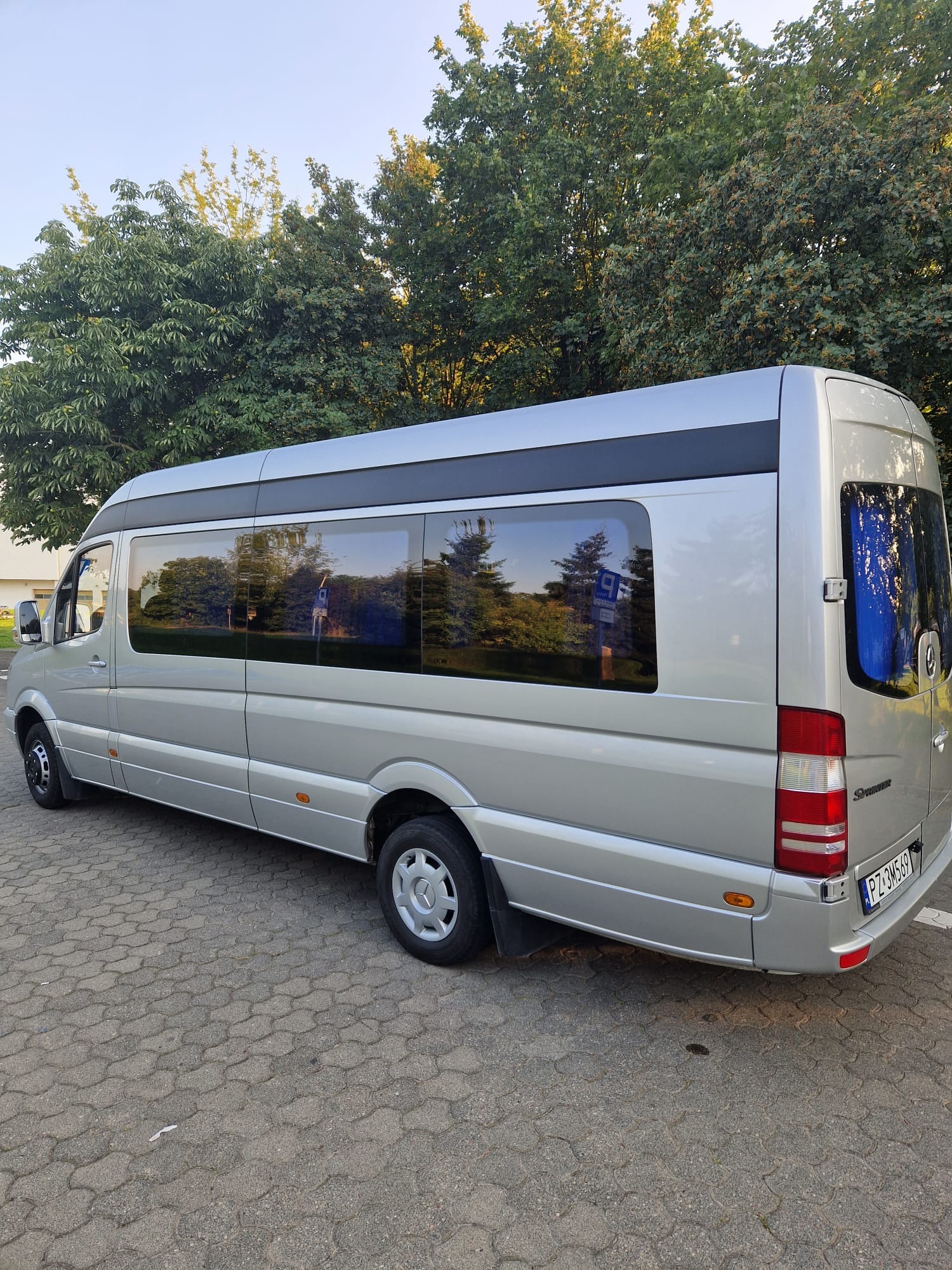 Srebrny bus Mercedes Sprinter z przyciemnianymi szybami na kostce brukowej, widok z boku i tyłu, z odbiciem drzew w oknach.