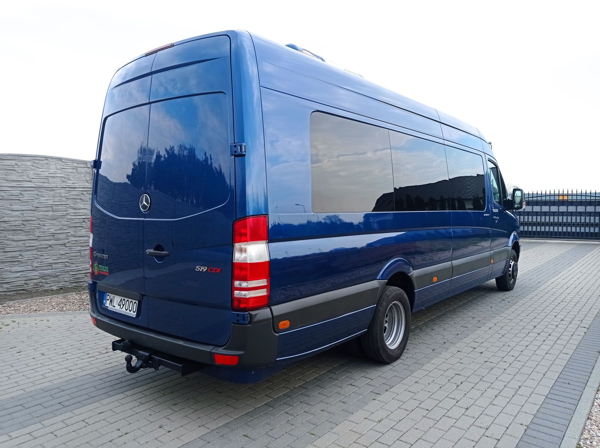 Niebieski bus Mercedes Sprinter 519 CDI z hakiem holowniczym, zaparkowany na brukowanym podjeździe, widok z tyłu.