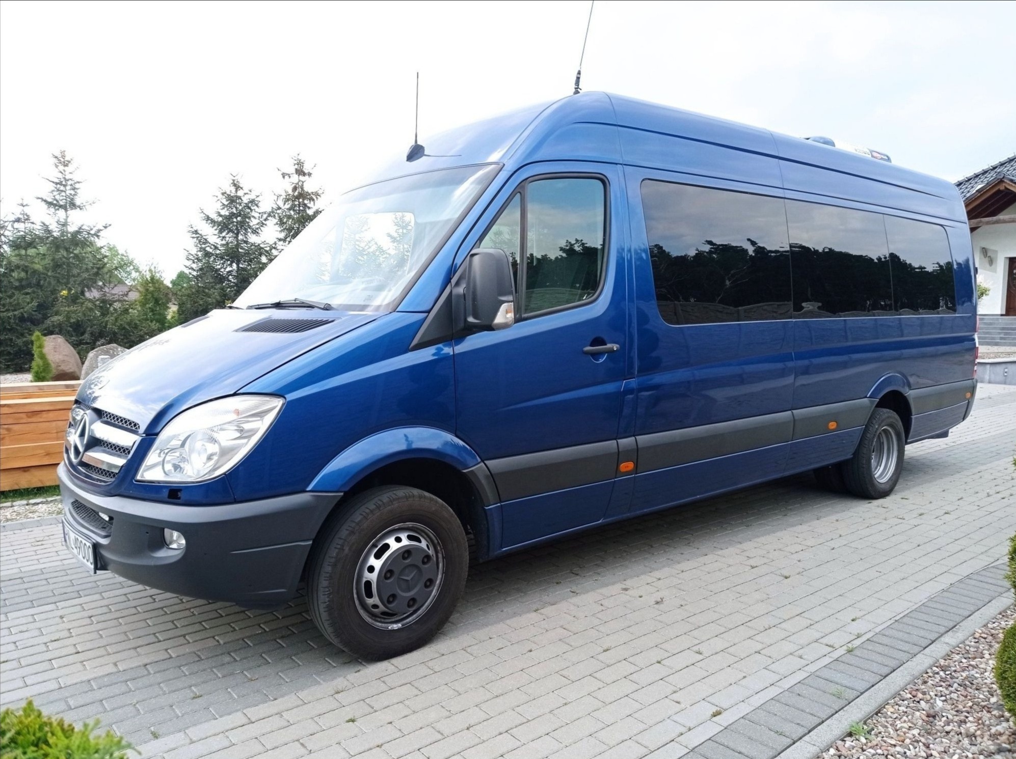 Niebieski bus Mercedes Sprinter z przyciemnianymi szybami, zaparkowany na kostce brukowej przed domem z ogrodem.