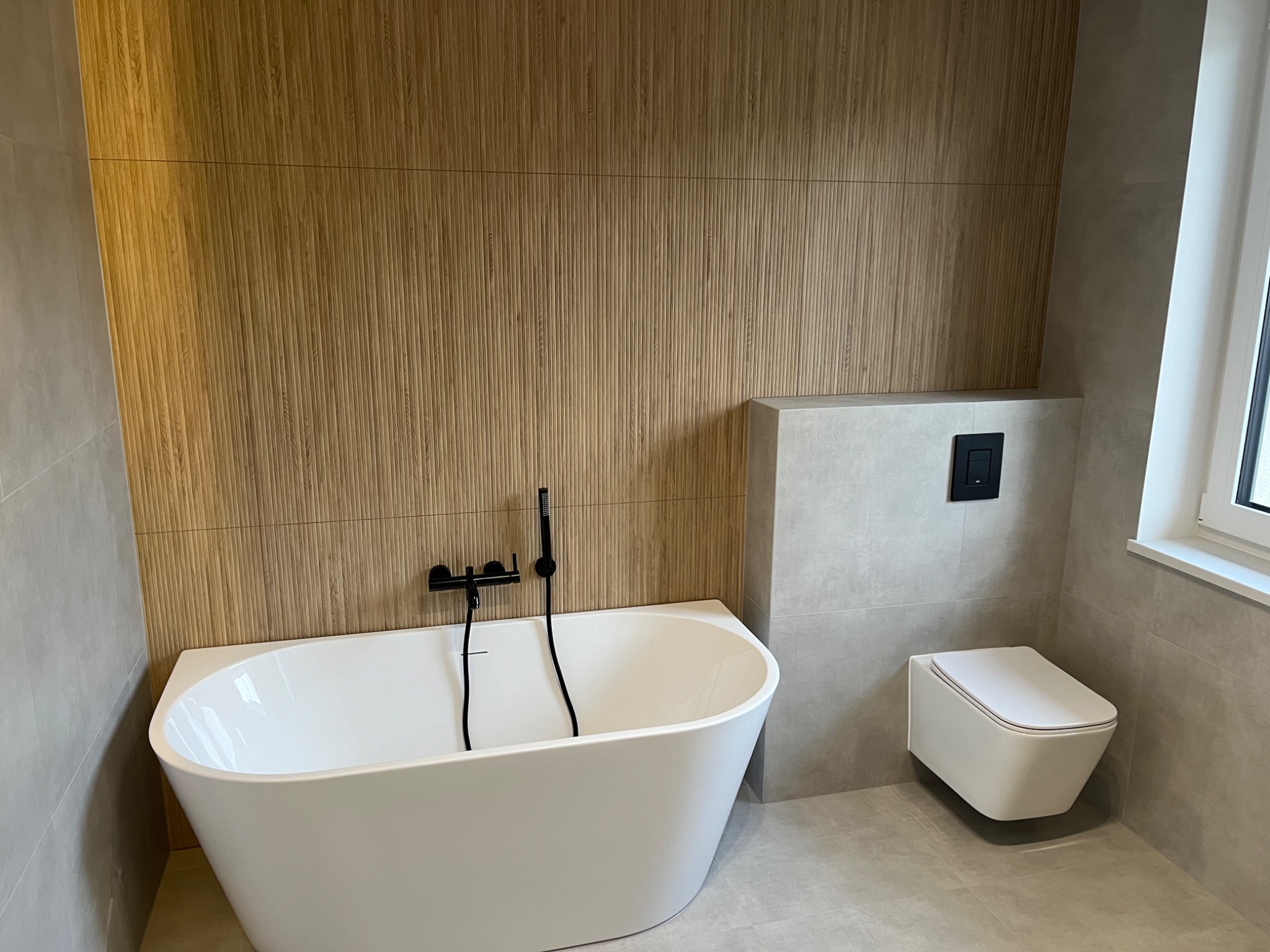 Nowoczesna łazienka z wanną wolnostojącą, drewnianą ścianą dekoracyjną i podwieszanym WC, wykończona szarymi płytkami.