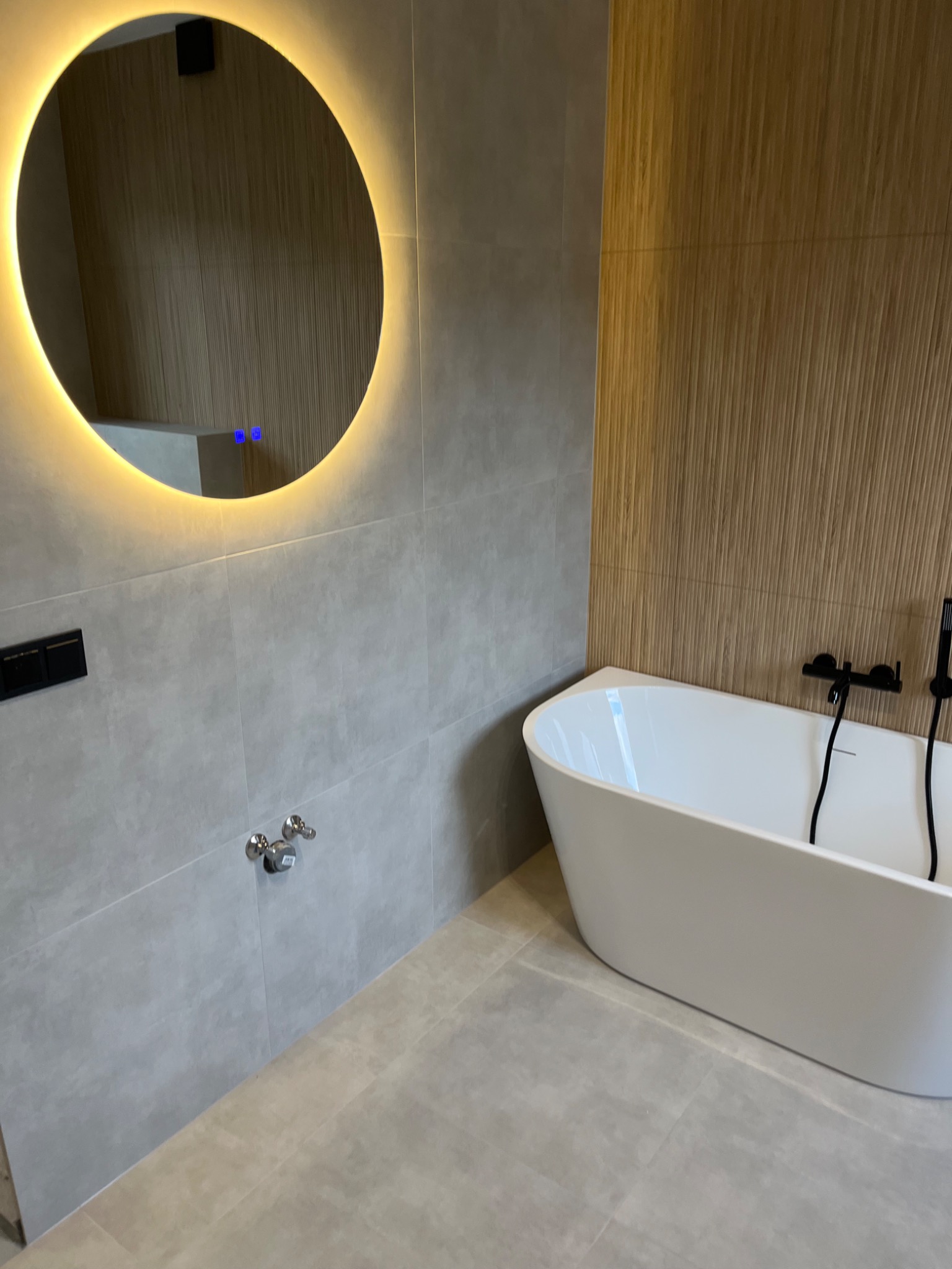 Nowoczesna łazienka z wolnostojącą białą wanną, okrągłym lustrem z podświetleniem LED, szarą płytką ścienną i podłogową oraz drewnianym panelem ściennym.