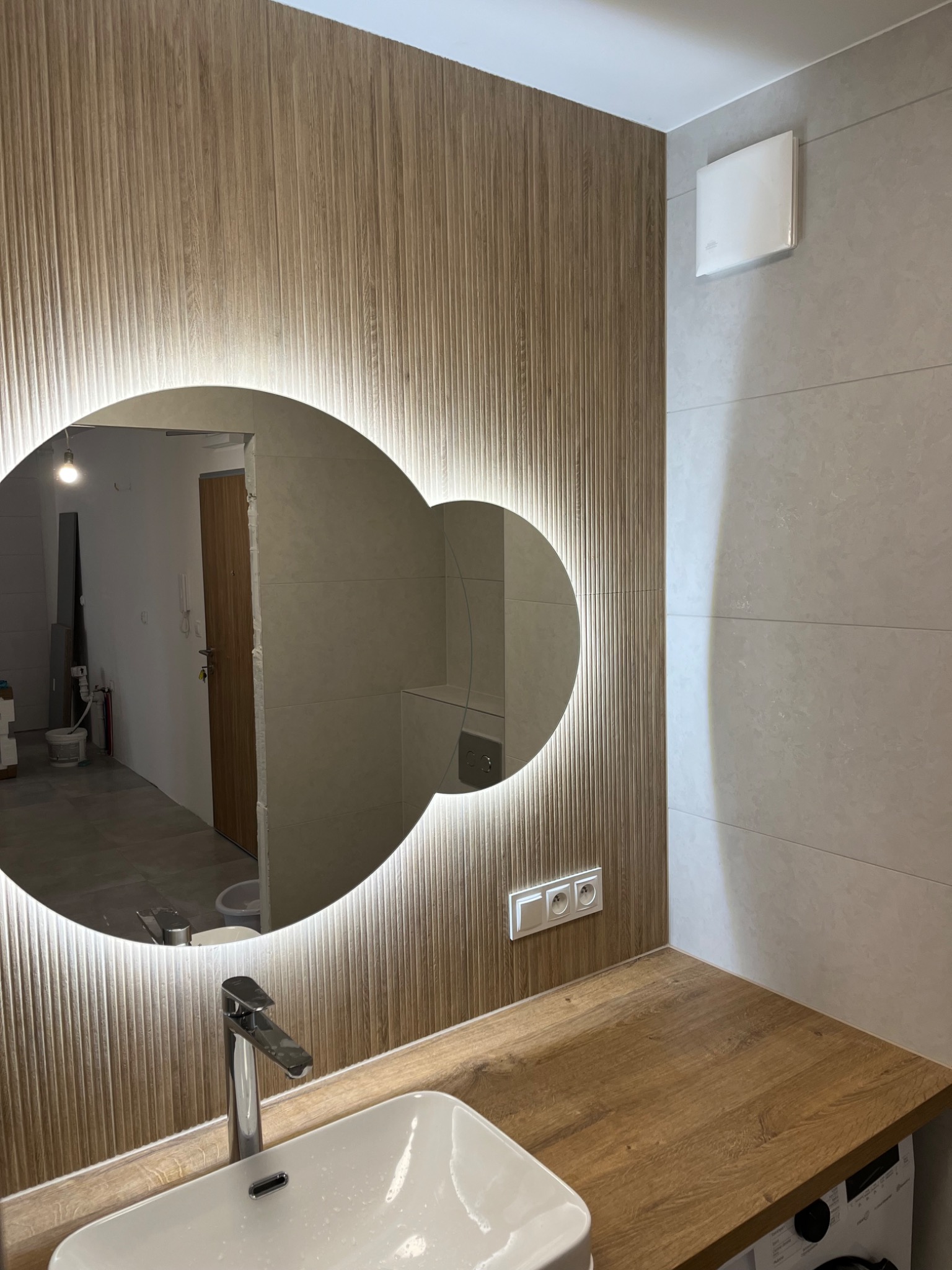 Wykończona łazienka z okrągłym lustrem podświetlanym LED, drewnopodobną ścianą z pionowymi żłobieniami, białą umywalką i baterią. W tle widoczne płytki oraz fragment wnętrza mieszkania w trakcie...