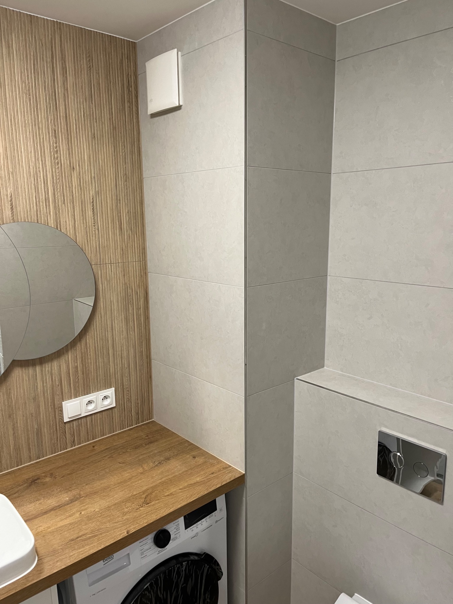 Nowoczesna łazienka po remoncie: drewnopodobna okładzina ścienna z okrągłym lustrem, blat zintegrowany z pralką, jasnoszare płytki na ścianach i zabudowa WC z chromowanym przyciskiem.