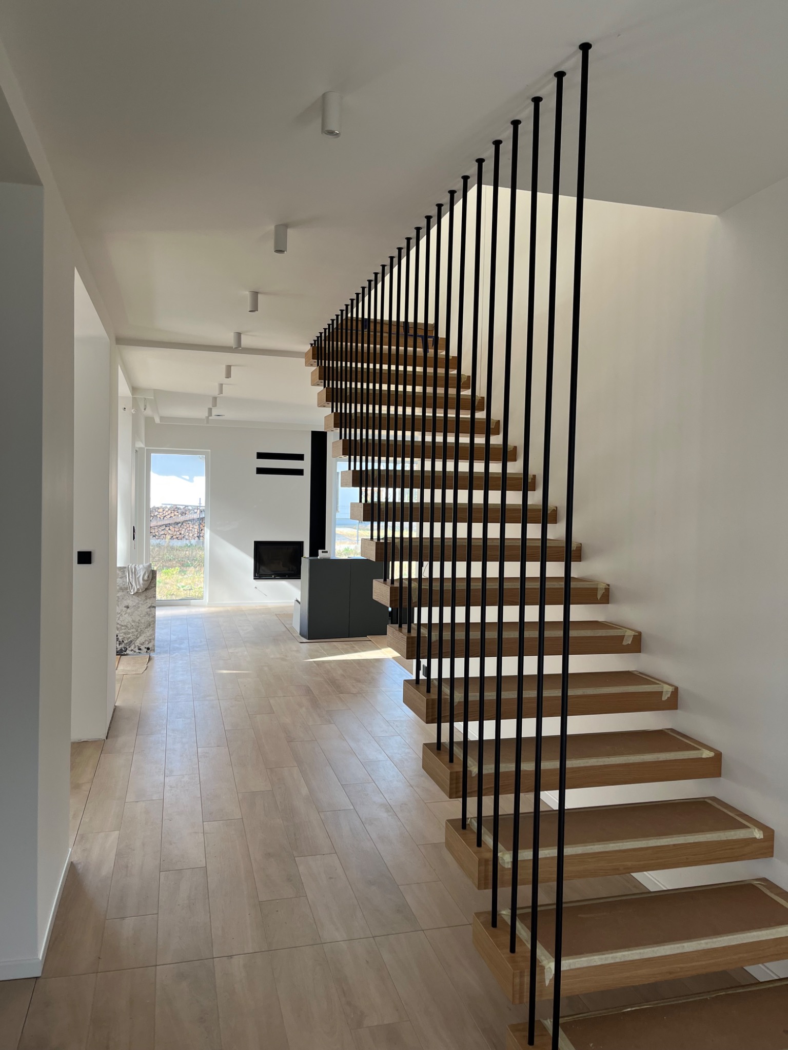 Wnętrze nowoczesnego domu z drewnianymi schodami o minimalistycznej konstrukcji z czarnymi, pionowymi szczeblami, zabezpieczonymi taśmą malarską na stopniach, widok na salon z kominkiem i okno...