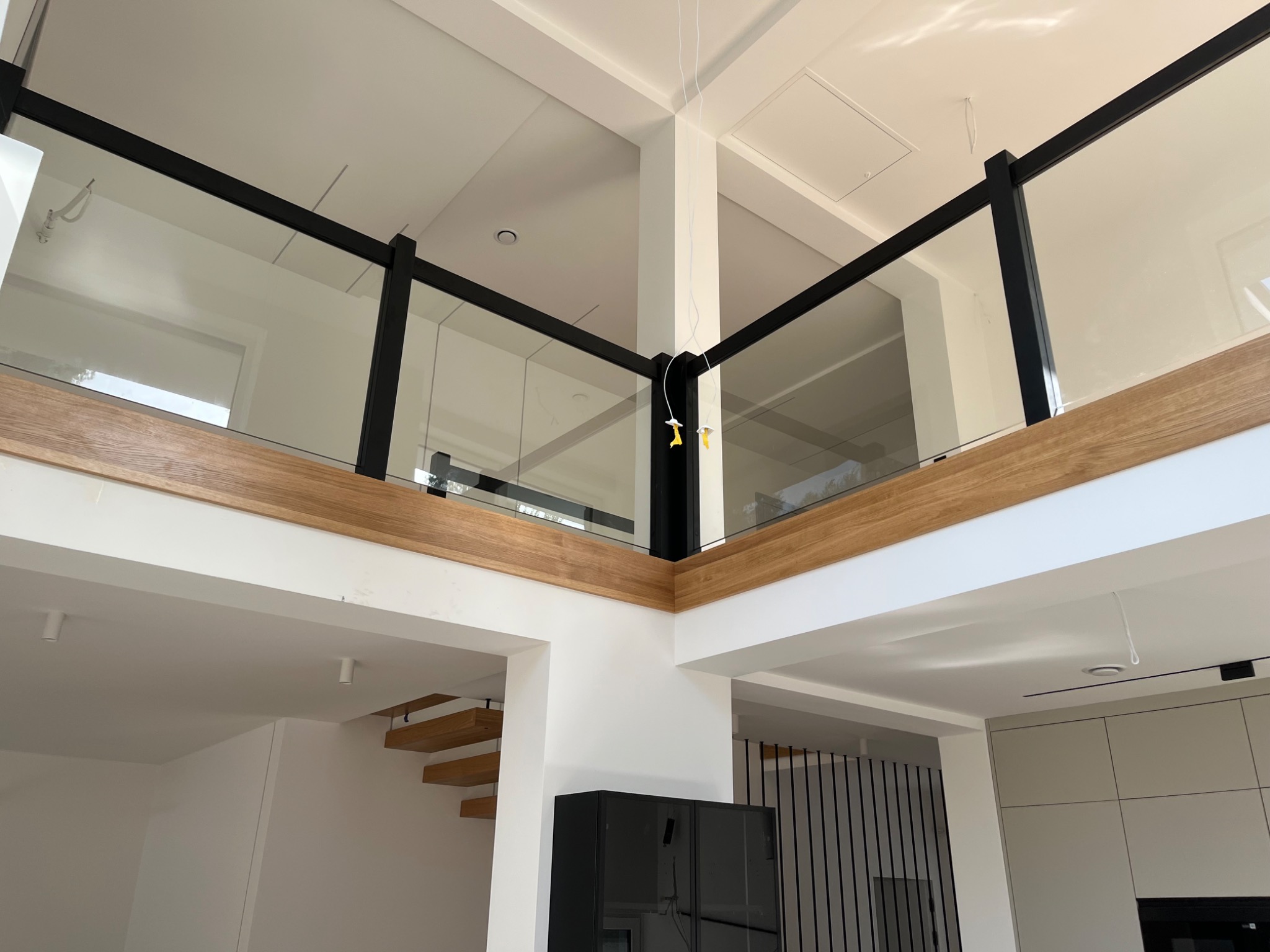 Wnętrze domu w stylu nowoczesnym z drewnianymi schodami i balustradą ze szkła z czarnymi elementami, widok z dołu na antresolę z oświetleniem punktowym.