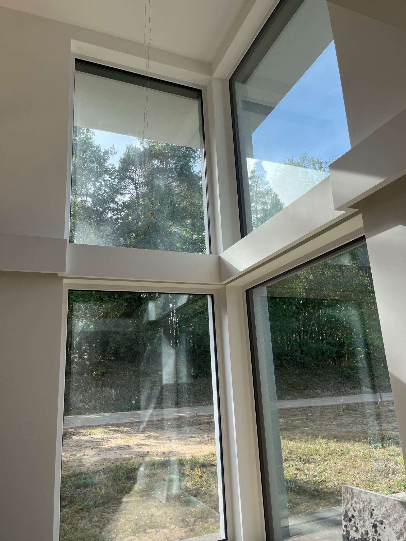 Wnętrze domu z dużymi, nowoczesnymi oknami z białymi ramami, widok na zewnątrz ukazuje drzewa i niezagospodarowany teren, roleta okienna częściowo zasłonięta.