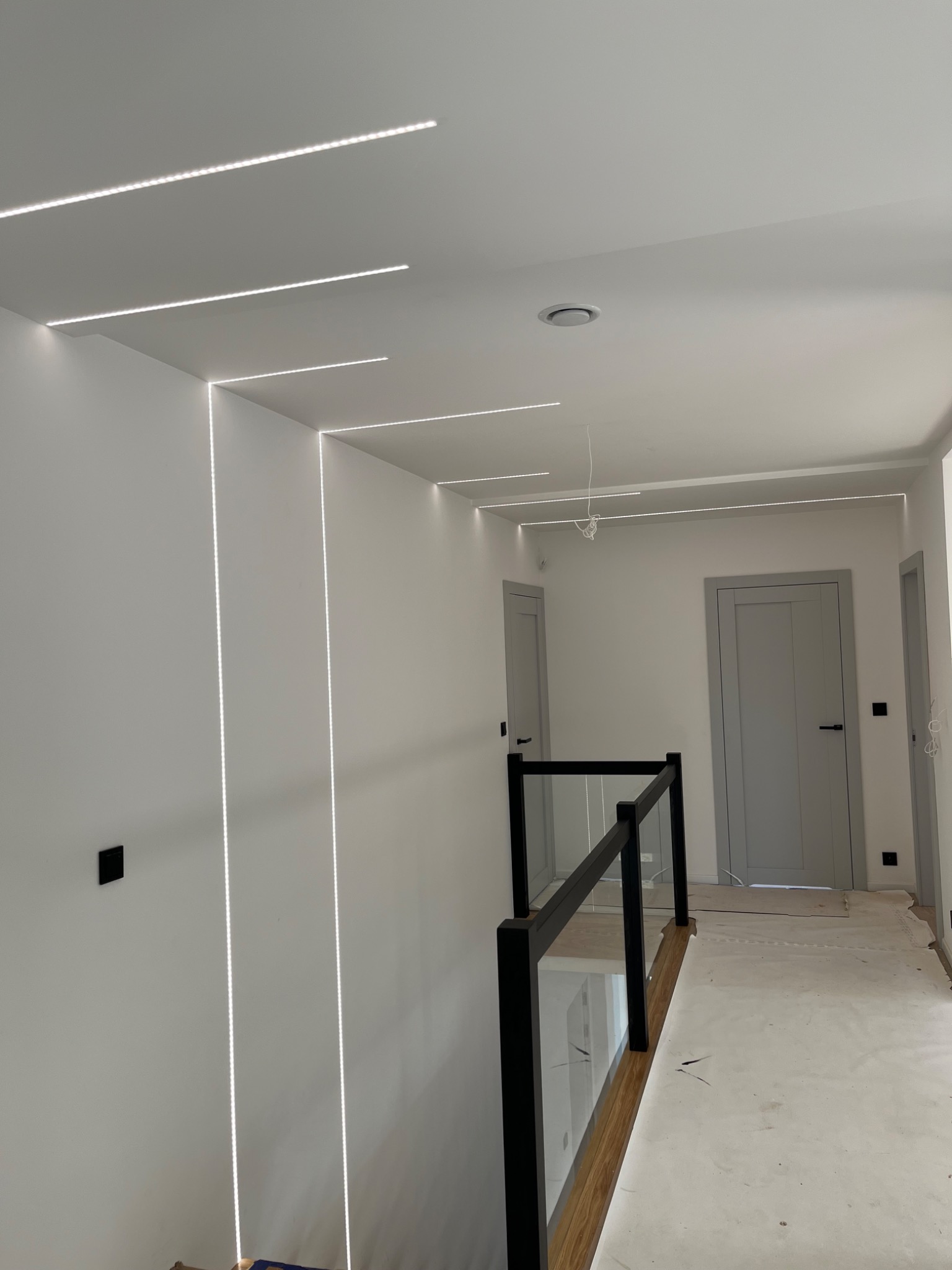 Nowoczesne oświetlenie LED w korytarzu z czarną balustradą i szarymi drzwiami, widoczne przewody na suficie przed zakończeniem montażu.