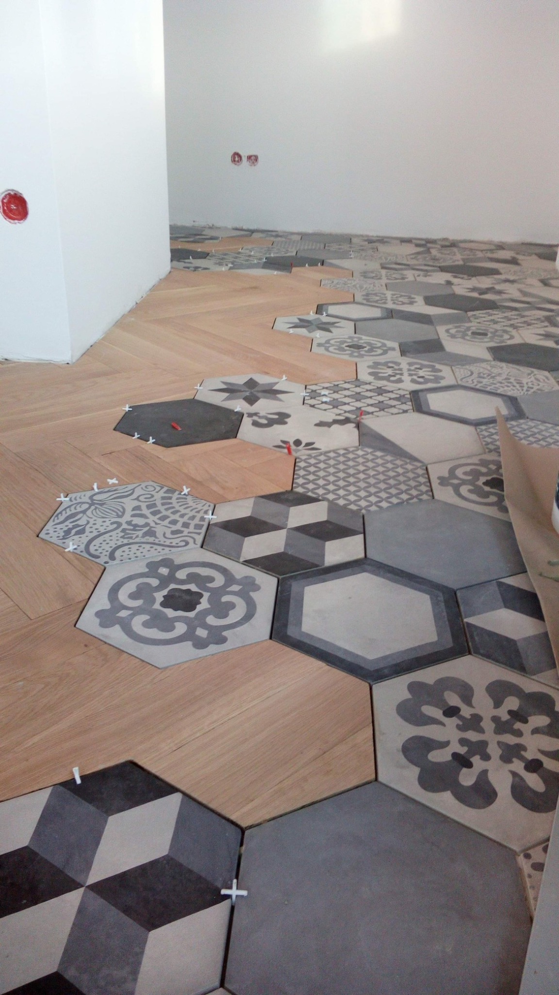 Układanie podłogi: heksagonalne płytki ceramiczne w odcieniach szarości z różnymi wzorami łączone z panelami ułożonymi w jodełkę, widoczne krzyżyki dystansowe i puszki elektryczne w ścianie.