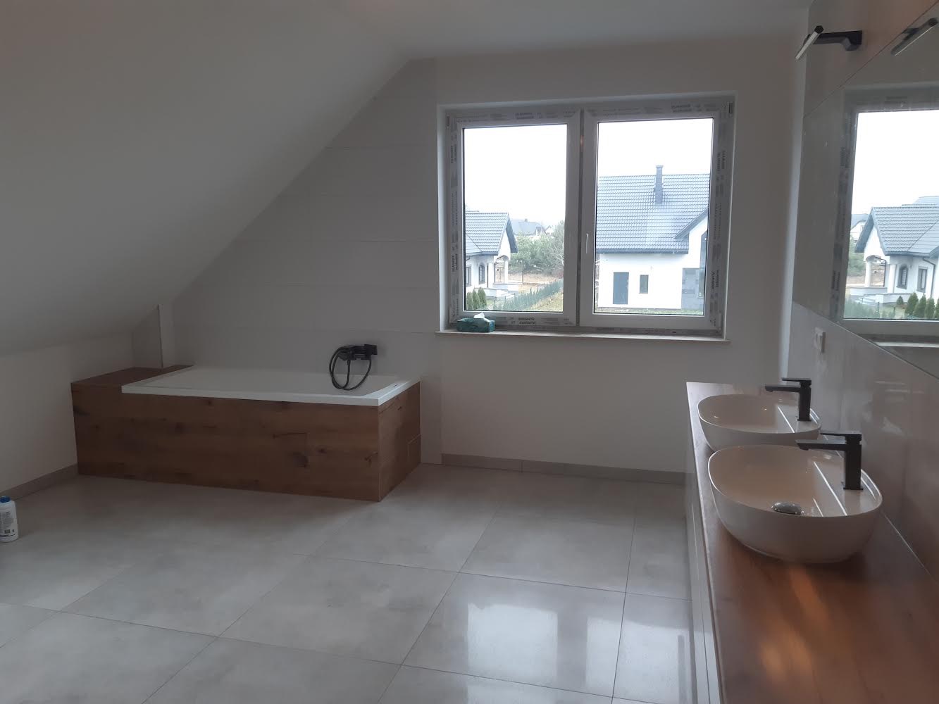 Nowoczesna łazienka na poddaszu z wanną obudowaną drewnem, dwiema białymi umywalkami na drewnianym blacie i szarymi płytkami podłogowymi, widok na domy za oknem.