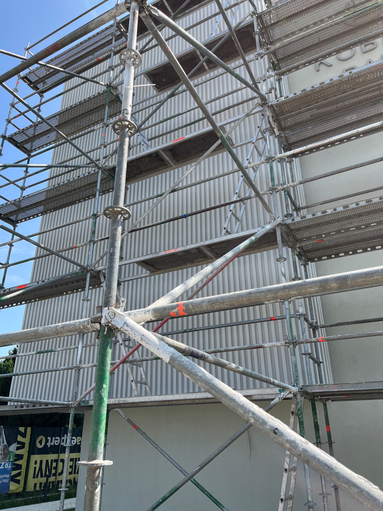 Budynek w trakcie prac elewacyjnych, widoczne rusztowania z metalowymi pomostami i pionowe panele elewacyjne w odcieniach szarości, częściowo otynkowana dolna partia ściany.