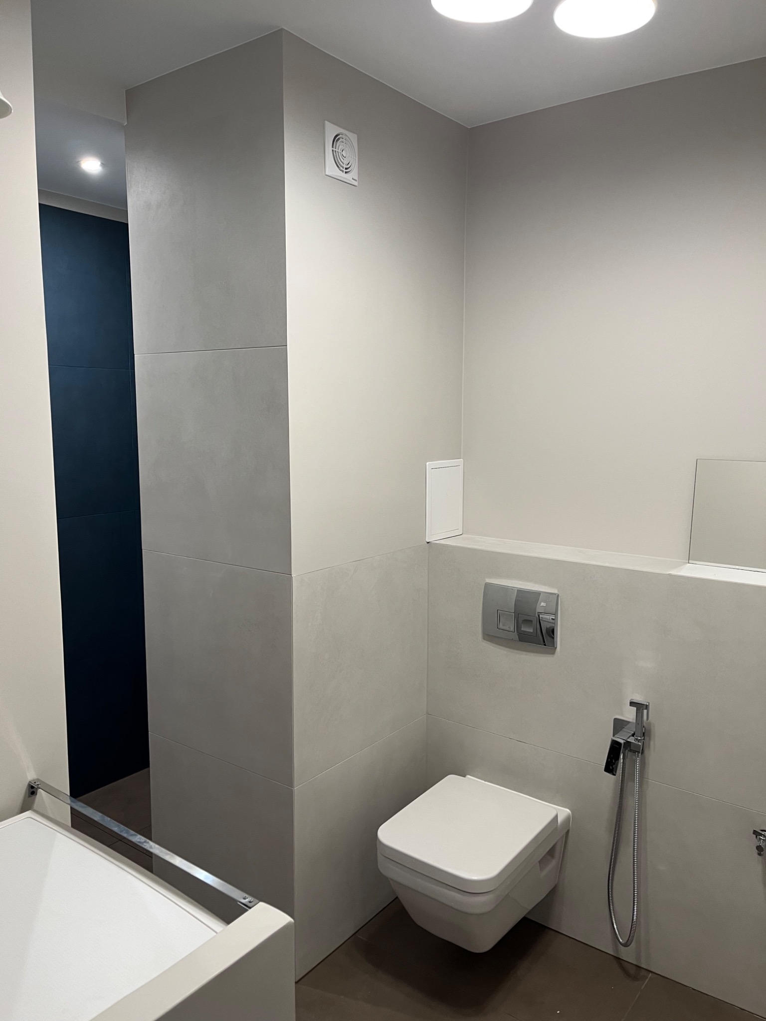 Nowoczesna, minimalistyczna łazienka z podwieszaną toaletą, szarą glazurą imitującą beton i ciemną podłogą. Widoczny chromowany zestaw bidetowy i biały właz rewizyjny.