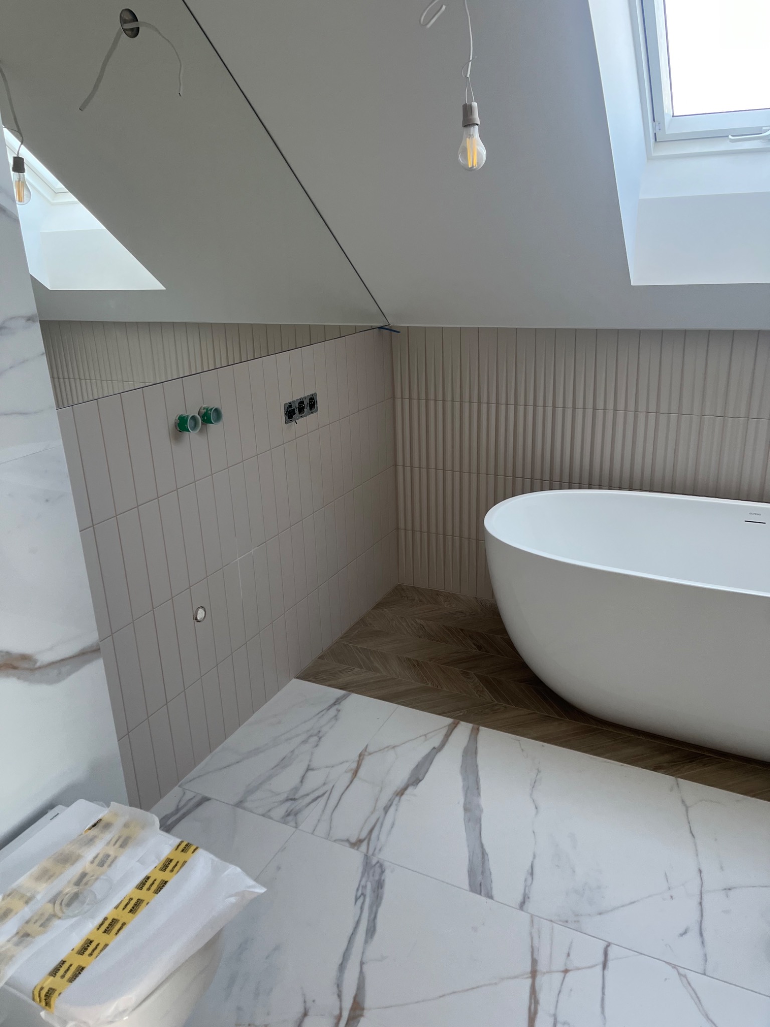 Nowoczesna łazienka w trakcie wykańczania: biała wolnostojąca wanna, marmurowe płytki podłogowe i ścienne, pionowe płytki na ścianie z instalacją wodną i elektryczną, drewnopodobna podłoga...