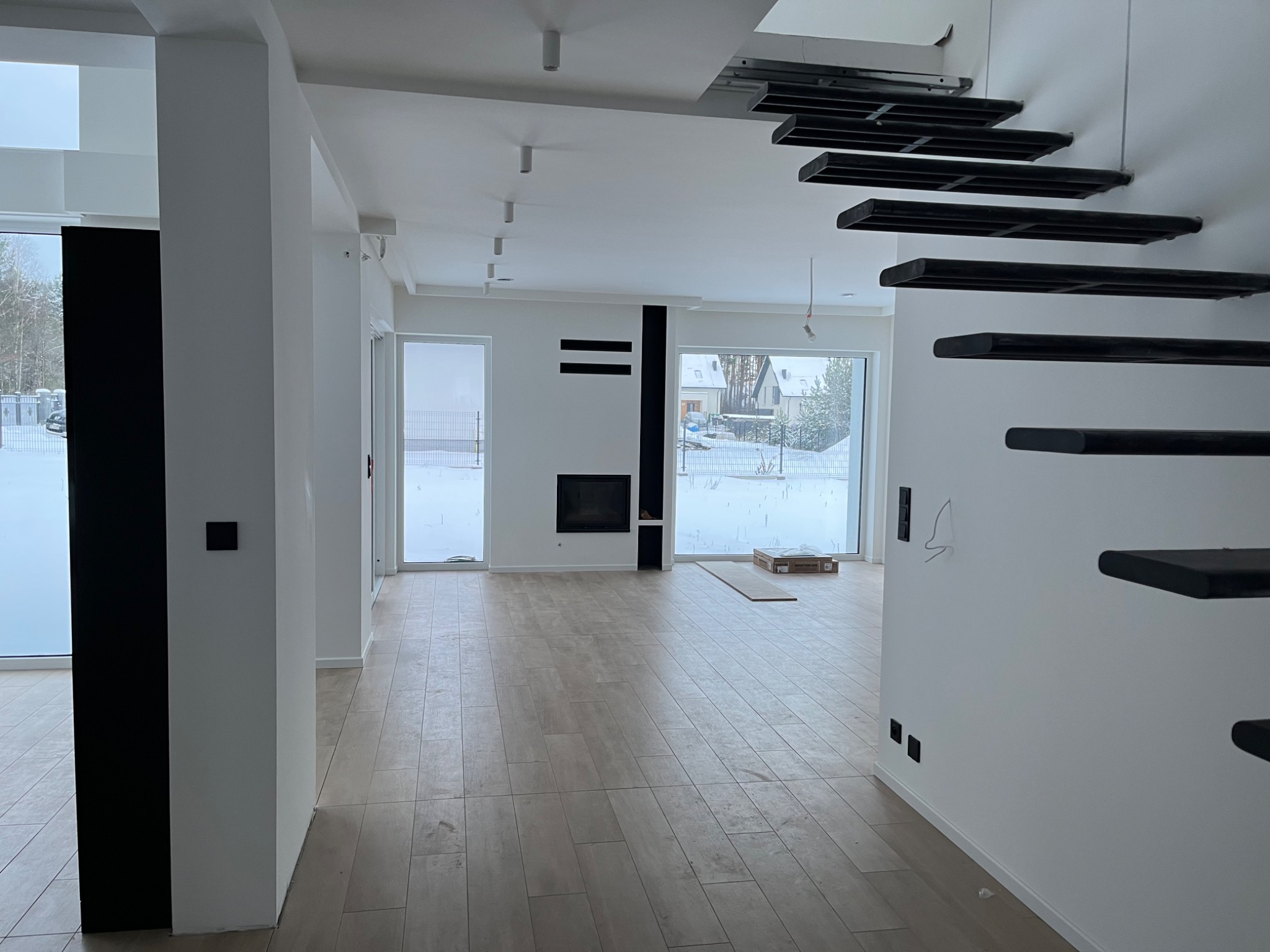 Jasne wnętrze domu w stylu minimalistycznym z drewnopodobną podłogą, czarnymi elementami dekoracyjnymi, nowoczesnym kominkiem i schodami konsolowymi; widoczne okna z zimowym krajobrazem na zewnątrz.