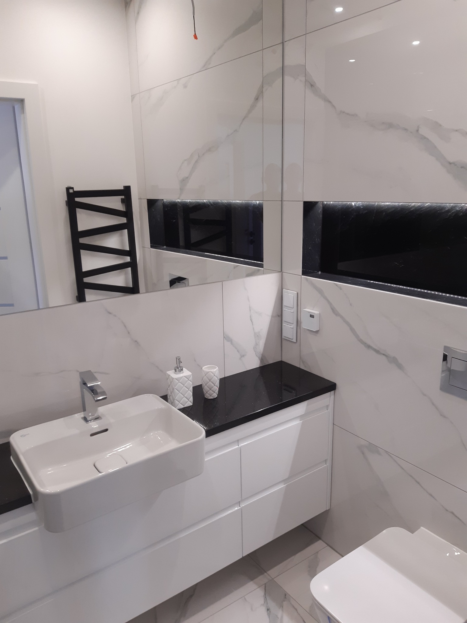 Nowoczesna łazienka z białą umywalką nablatową, armaturą chromowaną, grzejnikiem drabinkowym i płytkami imitującymi marmur na ścianach, z czarnym blatem i wnęką z podświetleniem LED.