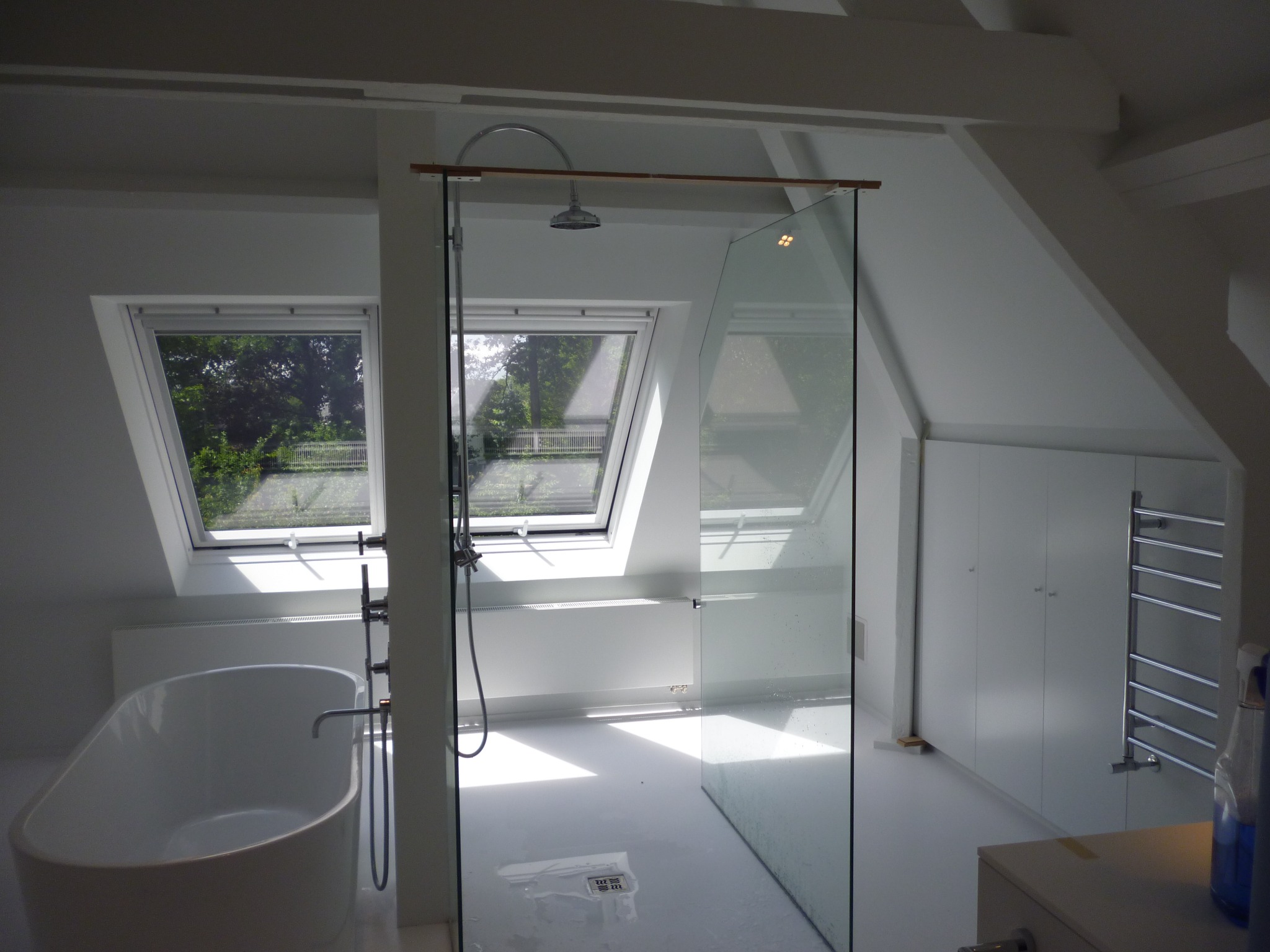 Jasna łazienka na poddaszu z oknami dachowymi, wolnostojącą wanną i kabiną prysznicową ze szklaną ścianką, widoczne grzejniki i szafki w zabudowie.