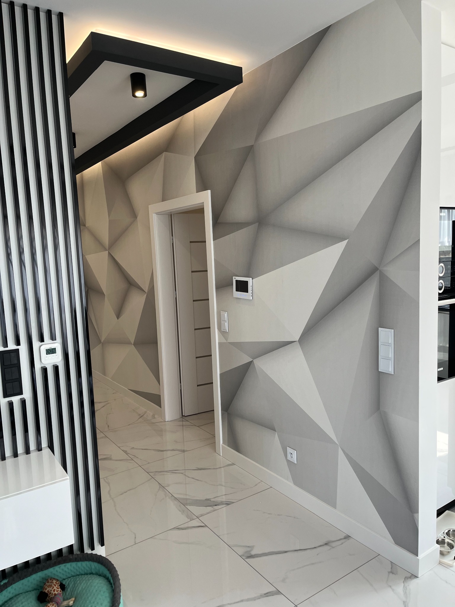 Nowoczesny przedpokój z geometryczną ścianą 3D w odcieniach szarości, białą podłogą z marmurkowym wzorem, czarnymi lamelami dekoracyjnymi i oświetleniem punktowym.