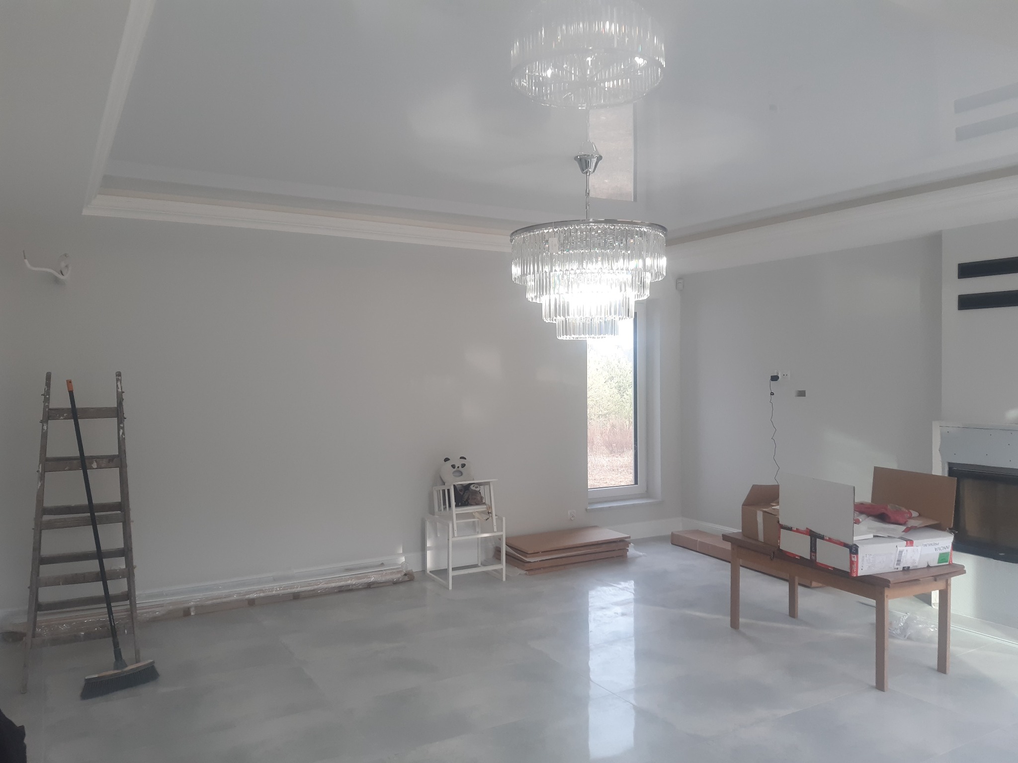 Wnętrze w trakcie prac wykończeniowych: szara podłoga, świeżo pomalowane ściany, drewniana drabina z miotłą oparta o ścianę, kryształowy żyrandol pod sufitem, kartony na stole, okno z widokiem...