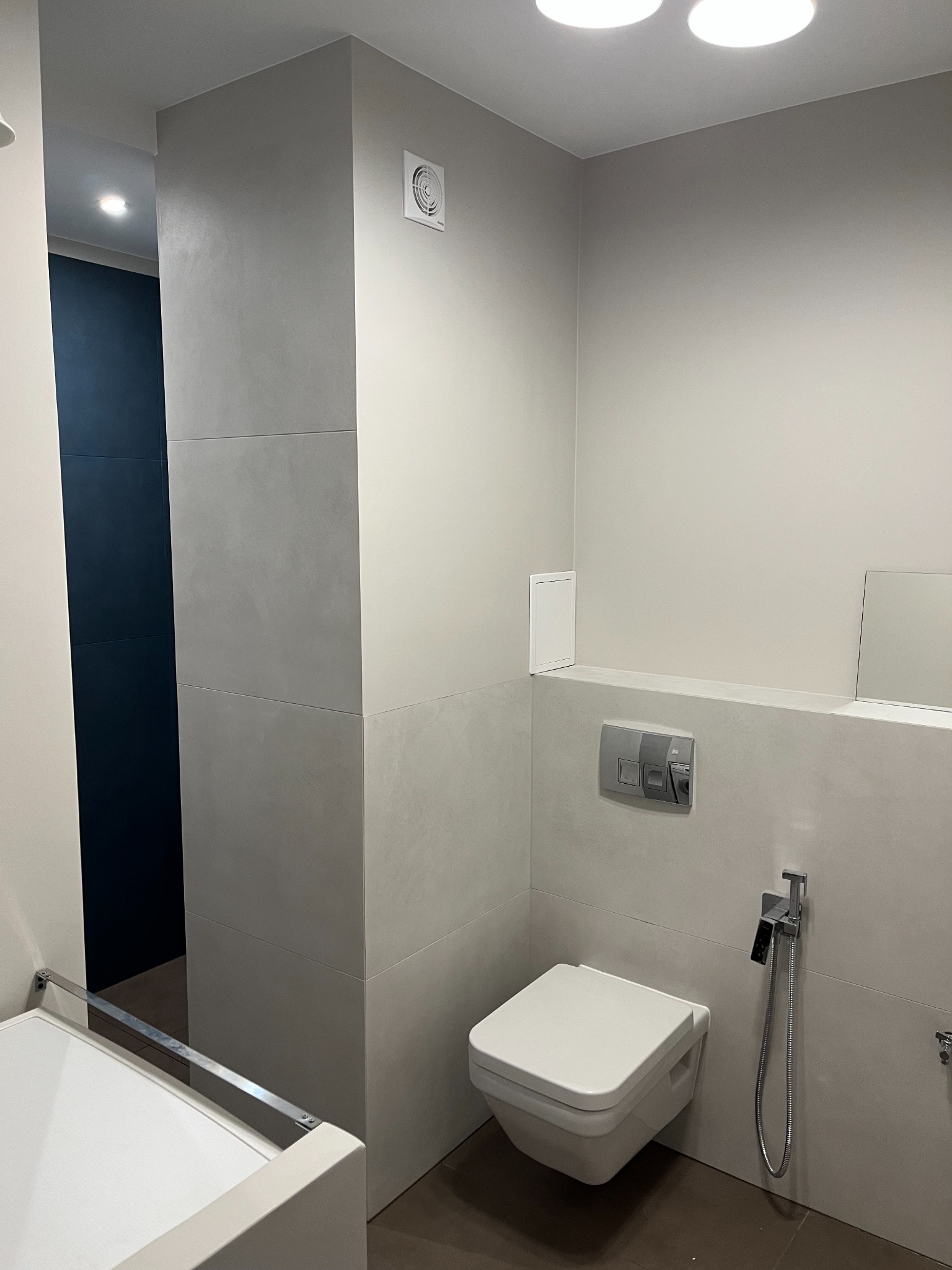 Nowoczesna łazienka z podwieszaną toaletą, szarą glazurą na ścianach i brązową podłogą, widoczny wentylator sufitowy i natrysk bidetowy.