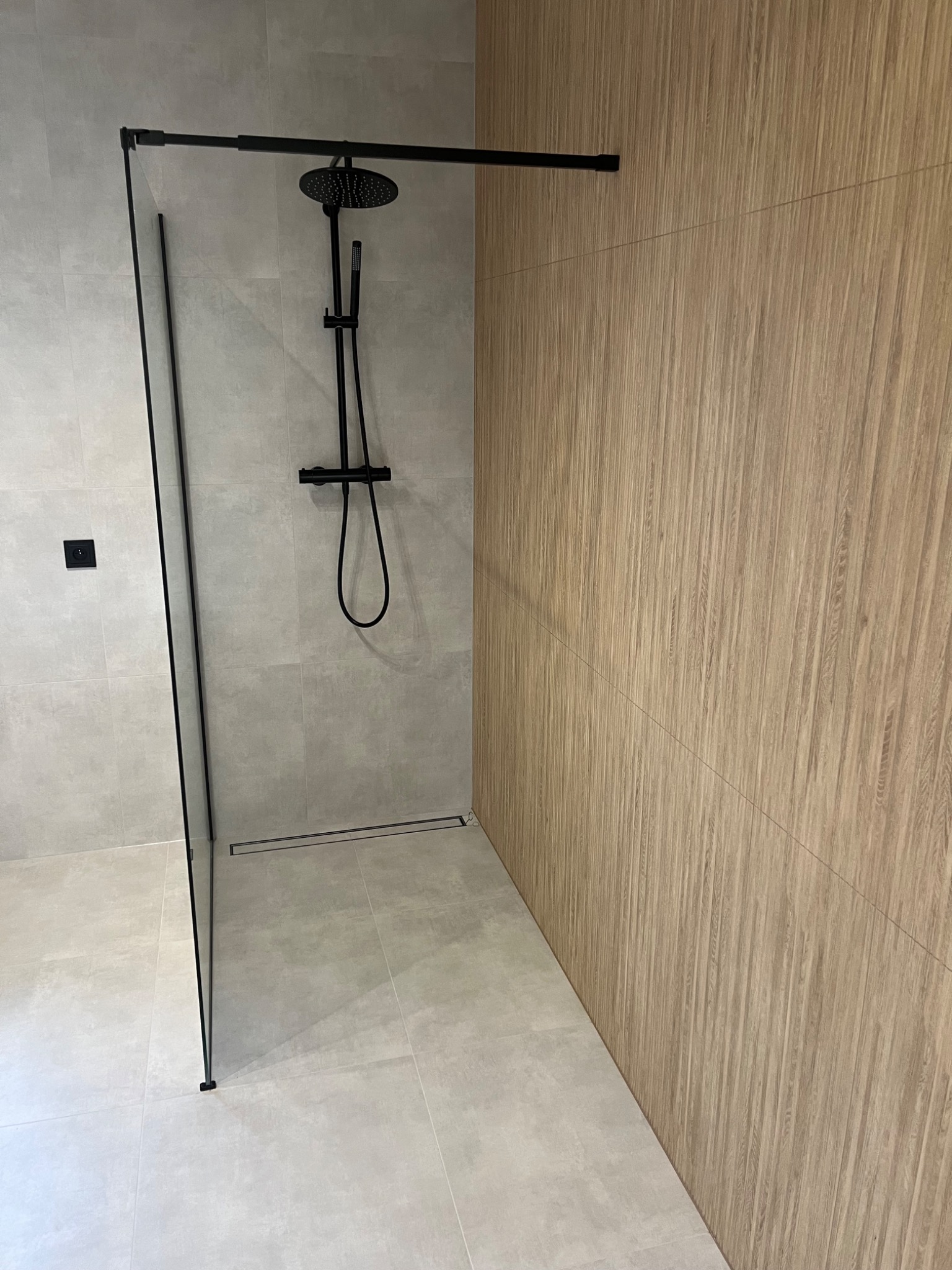 Nowoczesna kabina prysznicowa typu walk-in z czarną armaturą i odpływem liniowym, wykończona płytkami w odcieniach szarości i drewnopodobną okładziną ścienną.