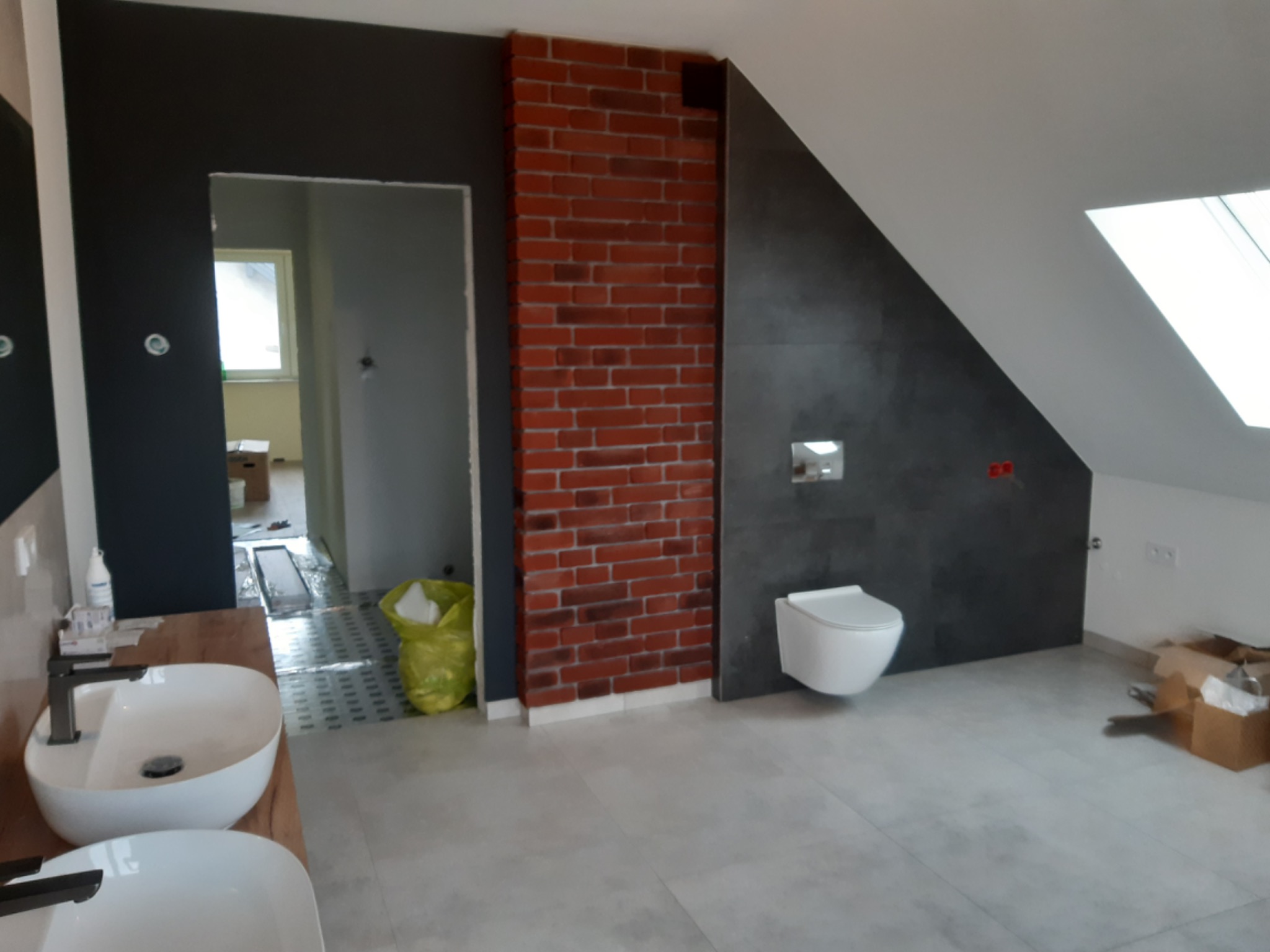 Wnętrze łazienki w trakcie remontu: widoczne dwie białe umywalki na drewnianym blacie, toaleta wisząca, fragment ściany z cegły, szare płytki podłogowe i fragment poddasza ze skosem.