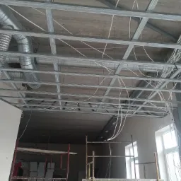 Montaż metalowej konstrukcji sufitu podwieszanego z widocznymi przewodami wentylacyjnymi i elektrycznymi na tle surowego betonowego stropu, z fragmentem ściany z płyt kartonowo-gipsowych...