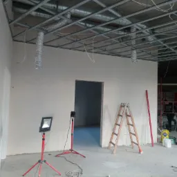 Wnętrze biura w trakcie prac wykończeniowych: ściany po szpachlowaniu, metalowa konstrukcja sufitu podwieszanego, oświetlenie robocze na statywach, drabina i materiały budowlane.