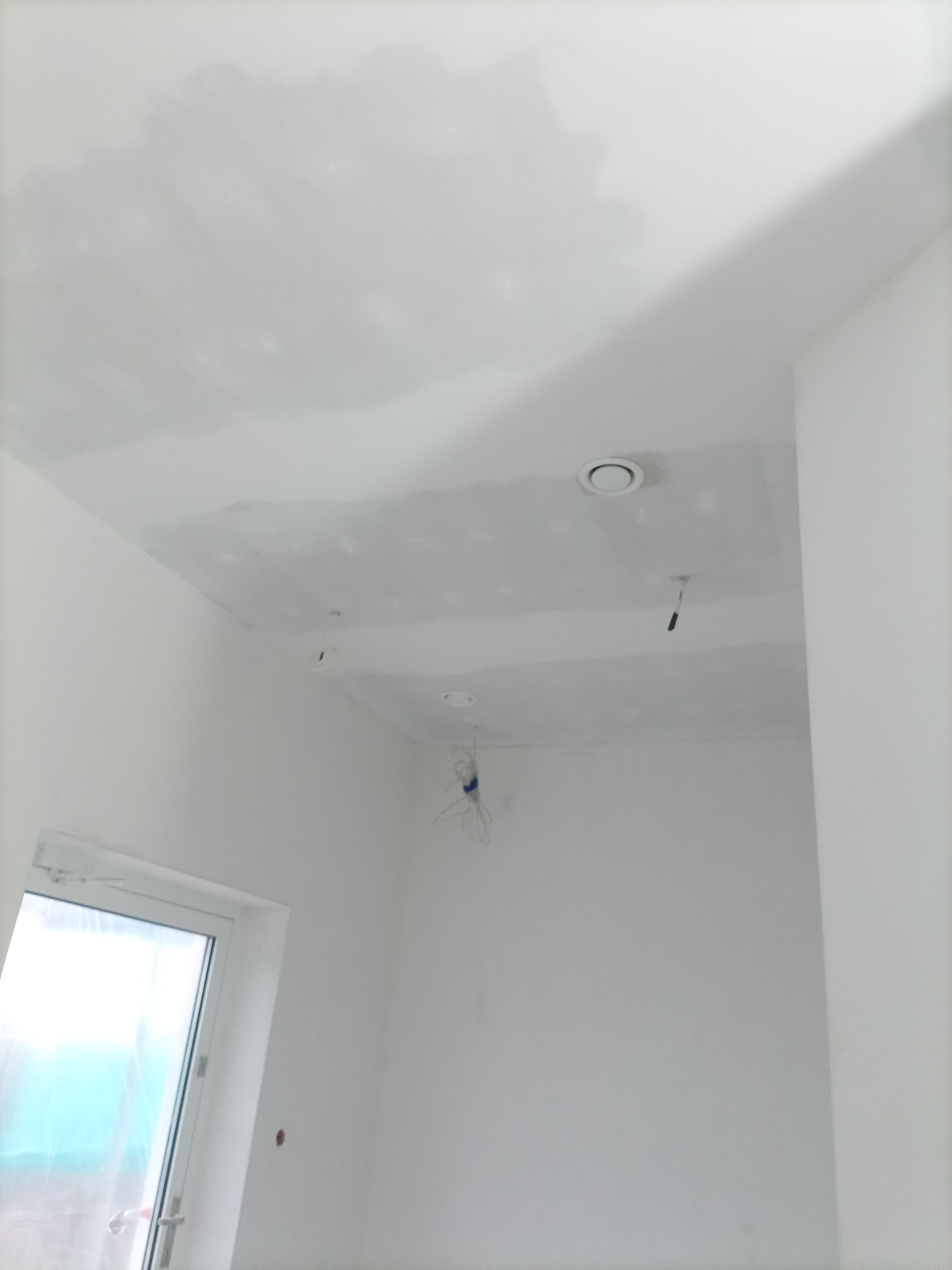 Sufit z płyt kartonowo-gipsowych w trakcie szpachlowania, widoczne otwory wentylacyjne i wystające przewody elektryczne, surowe wykończenie, okno w rogu pomieszczenia.