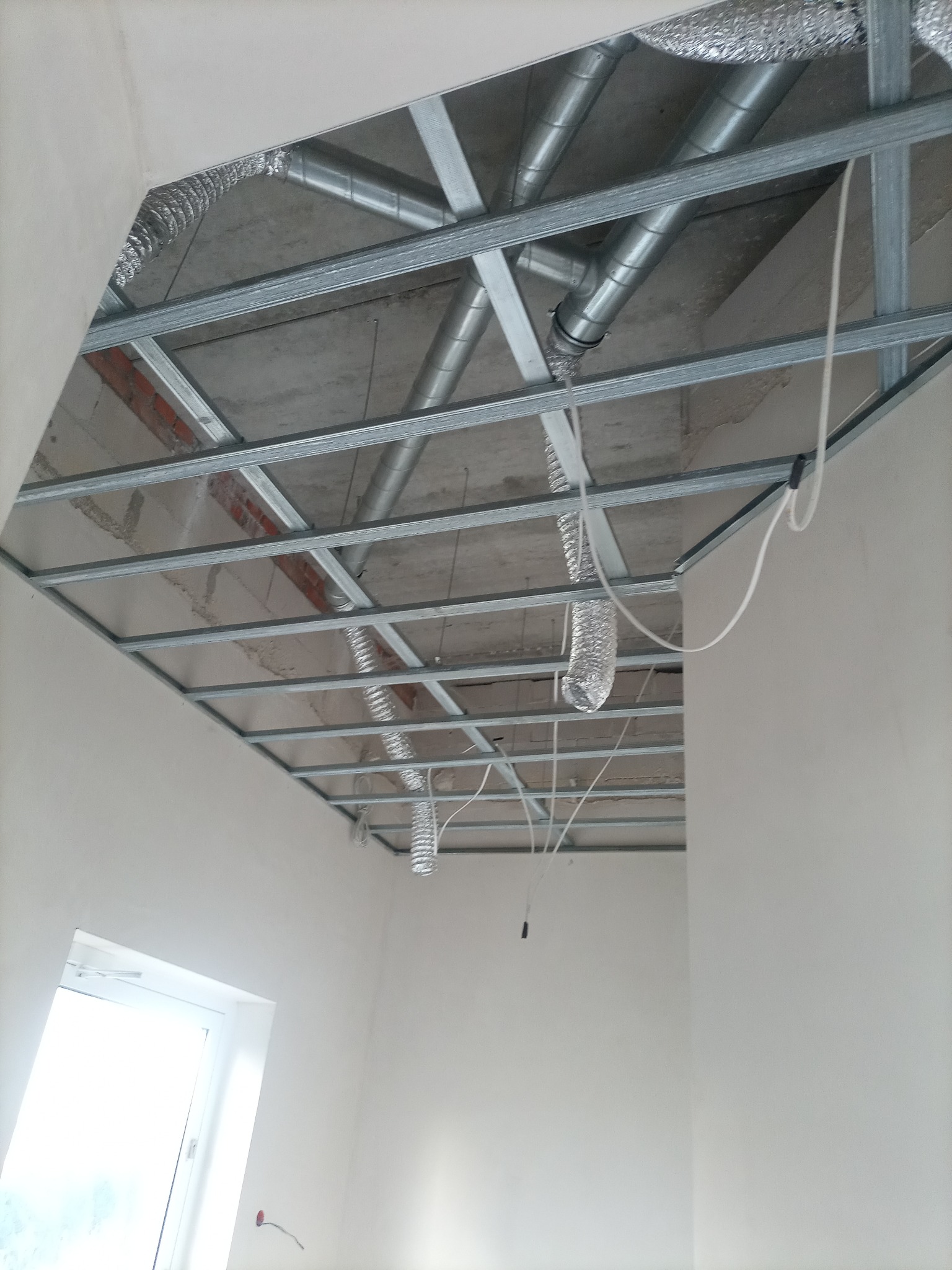 Metalowa konstrukcja sufitu podwieszanego z widocznymi elementami wentylacji w surowym stanie, białe ściany i okno w tle.