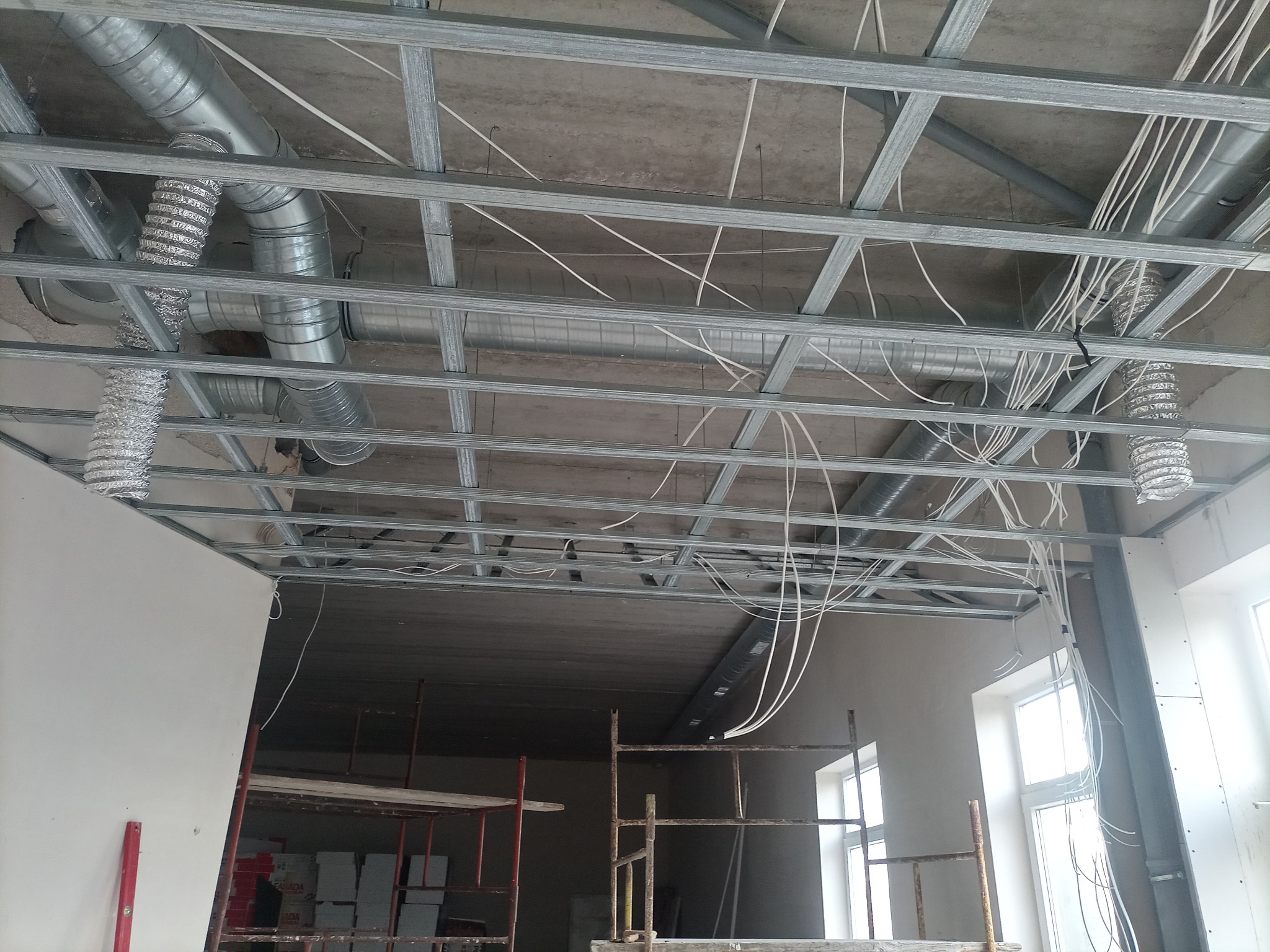 Montaż metalowej konstrukcji sufitu podwieszanego z widocznymi przewodami wentylacyjnymi i elektrycznymi na tle surowego betonowego stropu, z fragmentem ściany z płyt kartonowo-gipsowych...