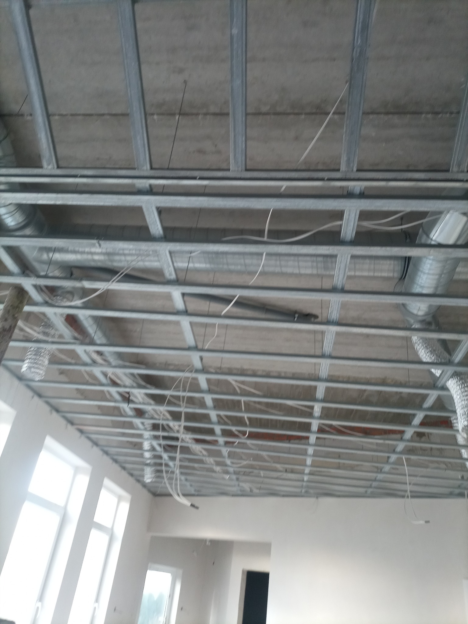 Metalowa konstrukcja sufitu podwieszanego z widocznymi przewodami wentylacyjnymi i kablami elektrycznymi, w tle jasne ściany i okna.