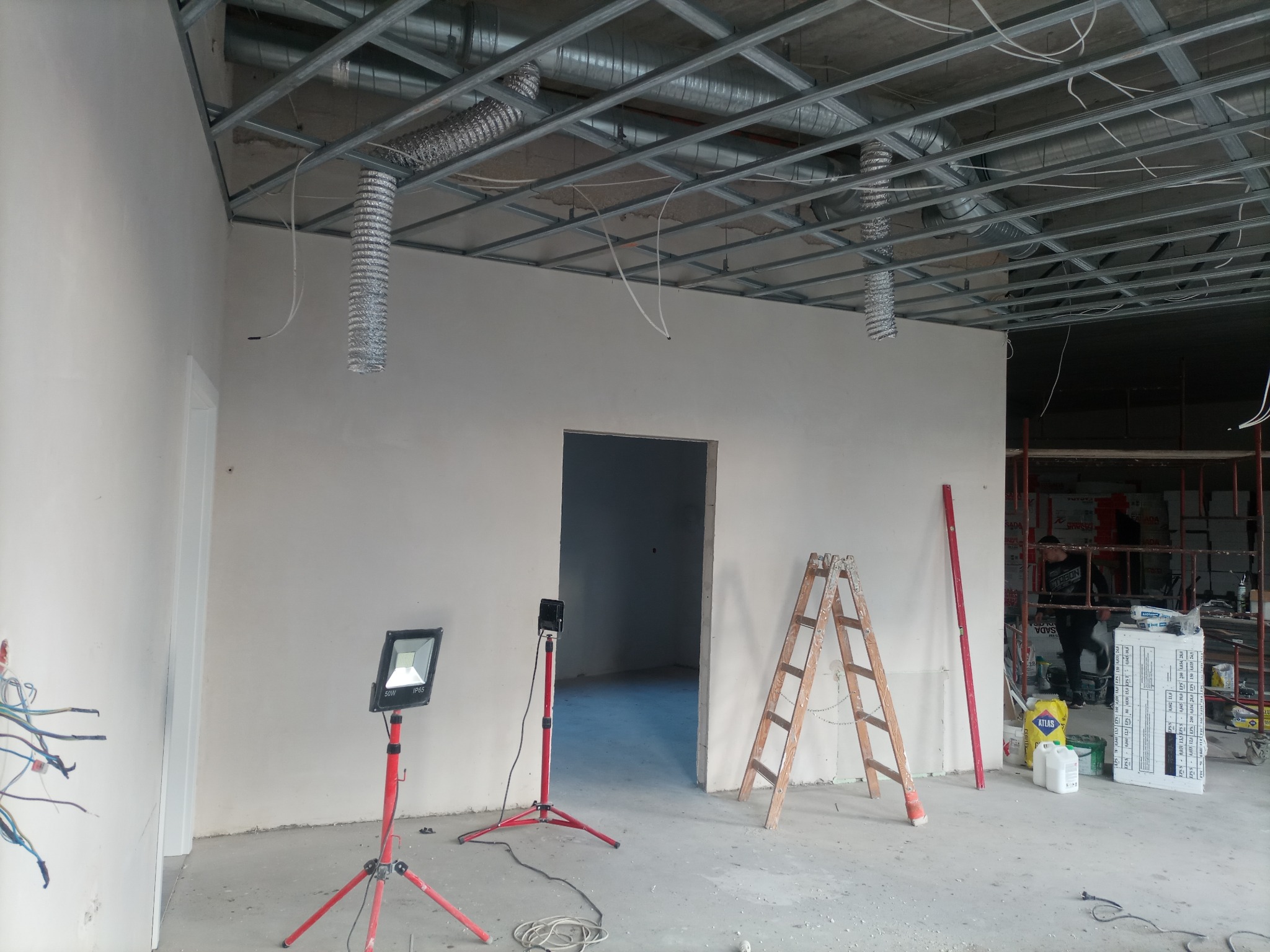 Wnętrze biura w trakcie prac wykończeniowych: ściany po szpachlowaniu, metalowa konstrukcja sufitu podwieszanego, oświetlenie robocze na statywach, drabina i materiały budowlane.