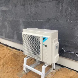 Zewnętrzna jednostka pompy ciepła Daikin Inverter R32, zamontowana na białym stelażu na podłożu z piasku, z widoczną czarną ścianą budynku z izolacją.