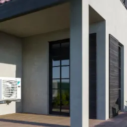 Biała jednostka zewnętrzna klimatyzatora z turkusowymi akcentami zamontowana na elewacji domu z drewnianym tarasem i meblami ogrodowymi w tle.