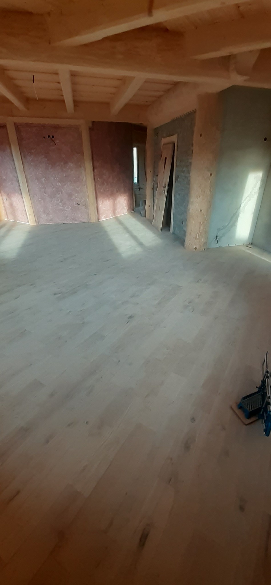 Jasne, drewniane panele podłogowe w surowym pomieszczeniu z drewnianym stropem i przygotowanymi ścianami do wykończenia, widoczny fragment narzędzia do cięcia paneli.