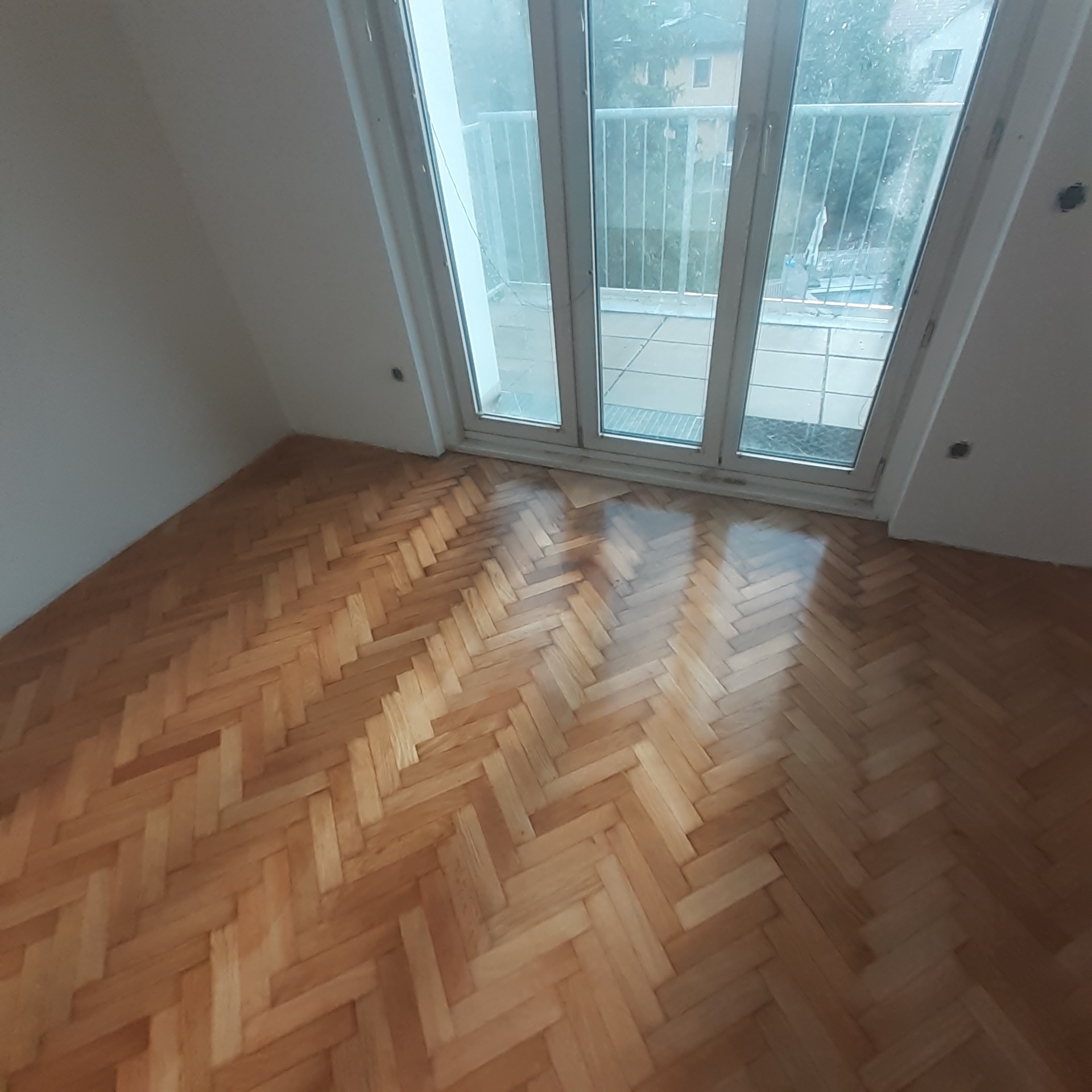 Ułożony parkiet w jodełkę w jasnym pokoju z dużym oknem balkonowym, widok na zewnątrz.